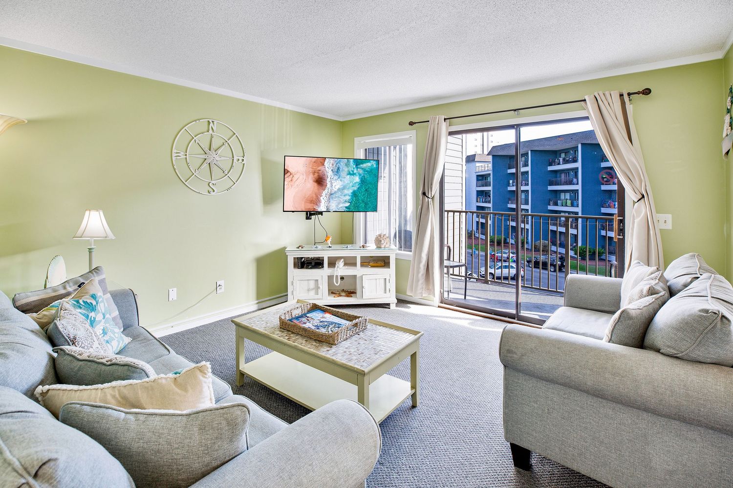 Bright living room with green walls, gray sofas, coffee table, and sliding glass doors opening to a balcony with ocean views.