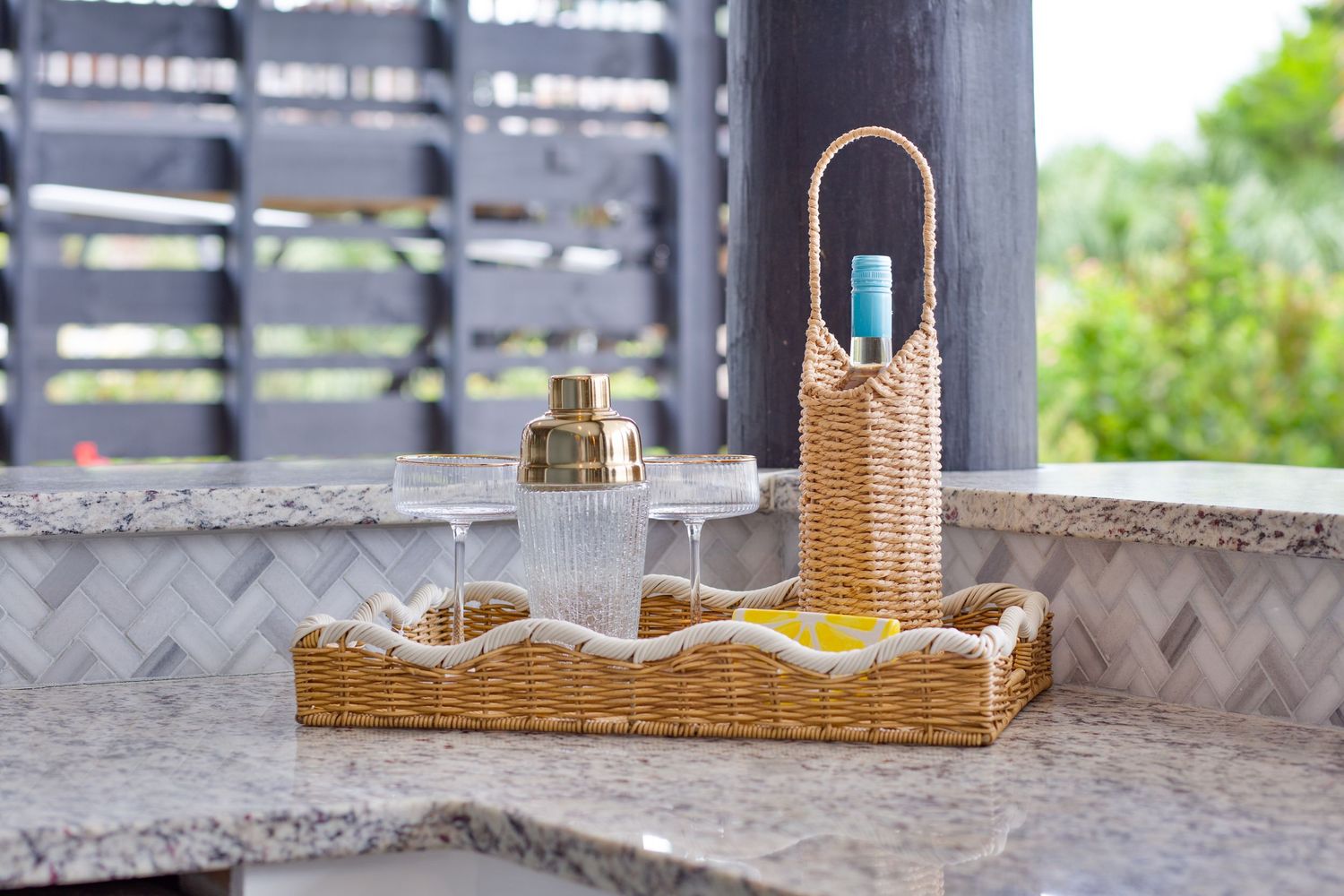 Wicker tray with cocktail shaker and glasses on granite countertop in outdoor bar area.