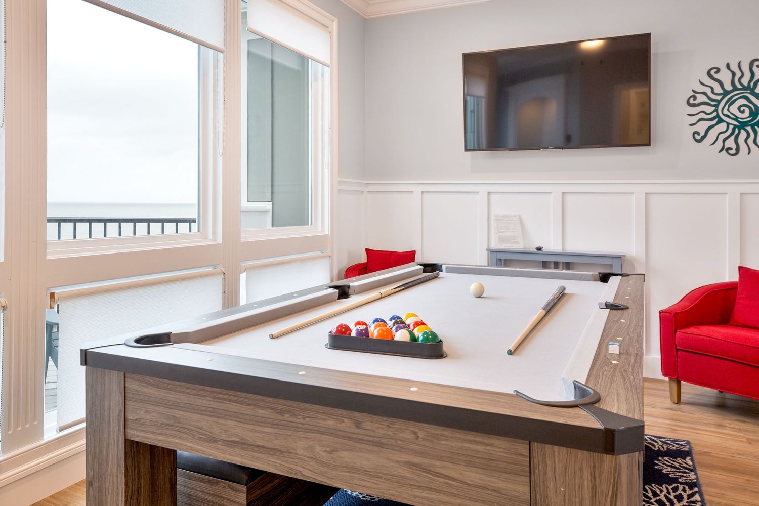 Game room with pool table, ocean view windows, wall-mounted TV, and red accent chairs.