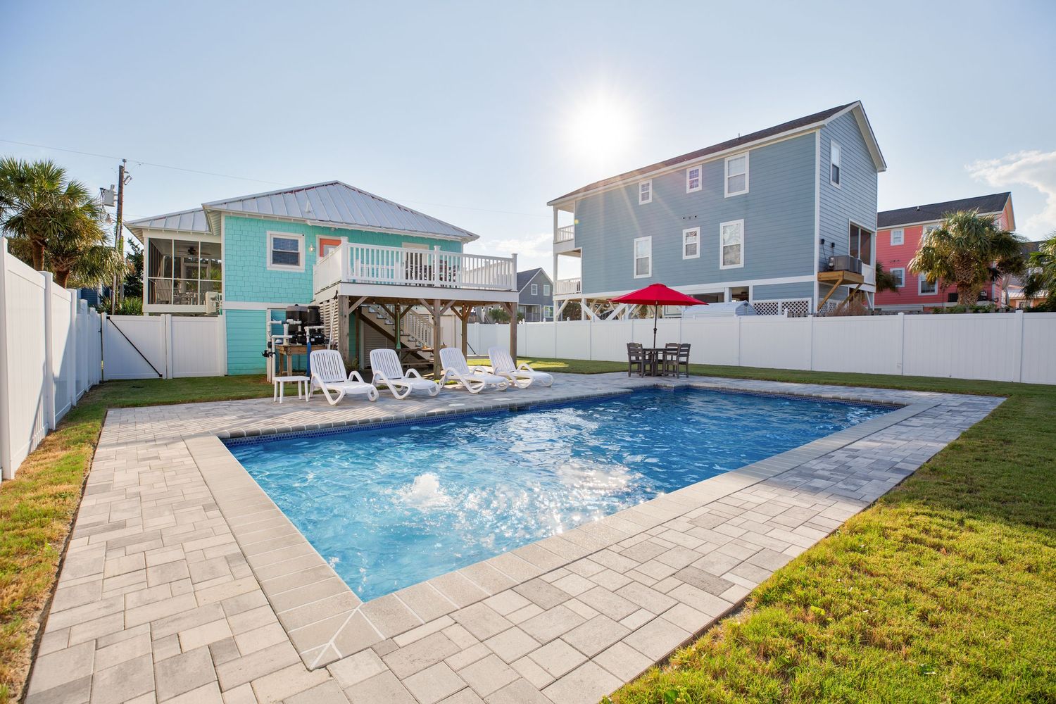 Bright sunny backyard with private pool, lounge chairs, and a colorful two-story beach house.