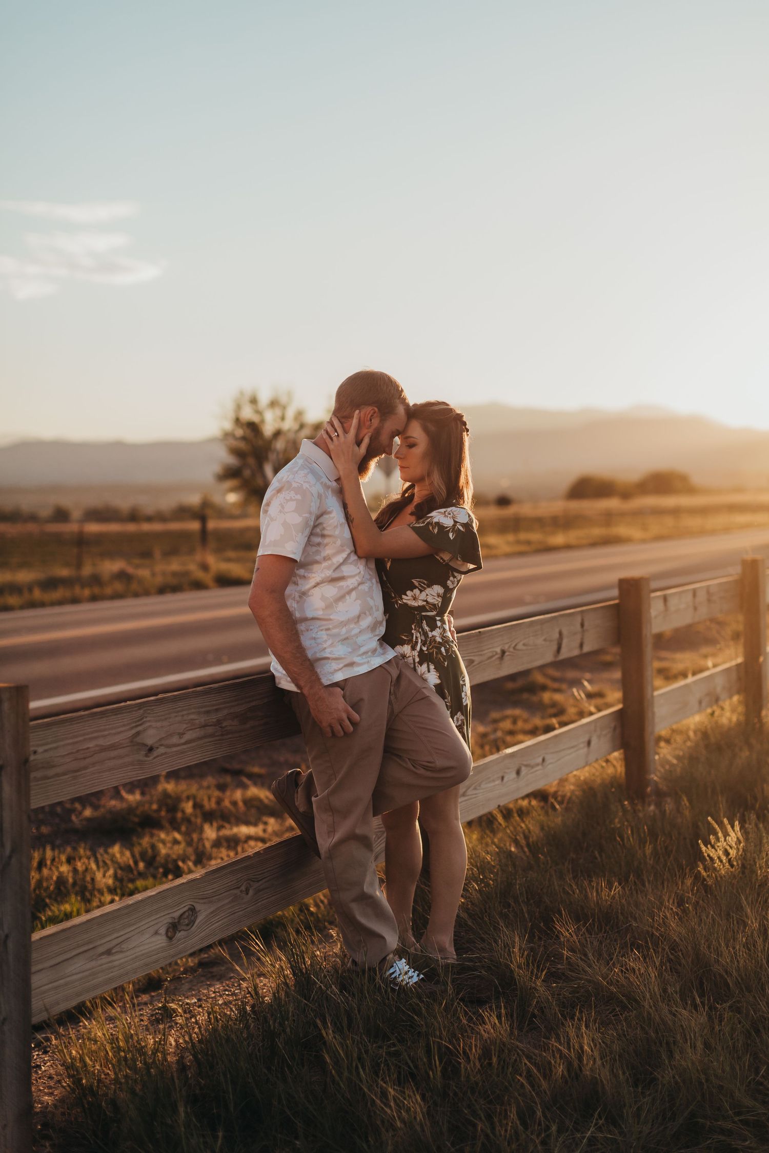 Romantic couple sharing an intimate moment against a wooden fence during golden hour sunset in a rural setting.