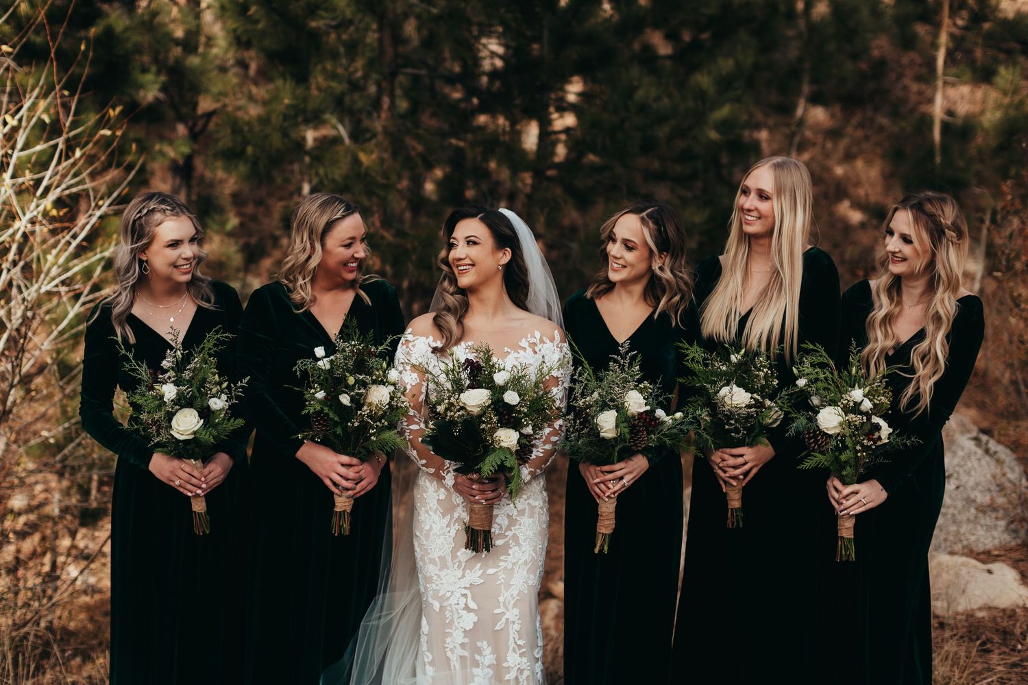 Bridal party in black dresses stands together holding white and green bouquets against an autumn forest backdrop.