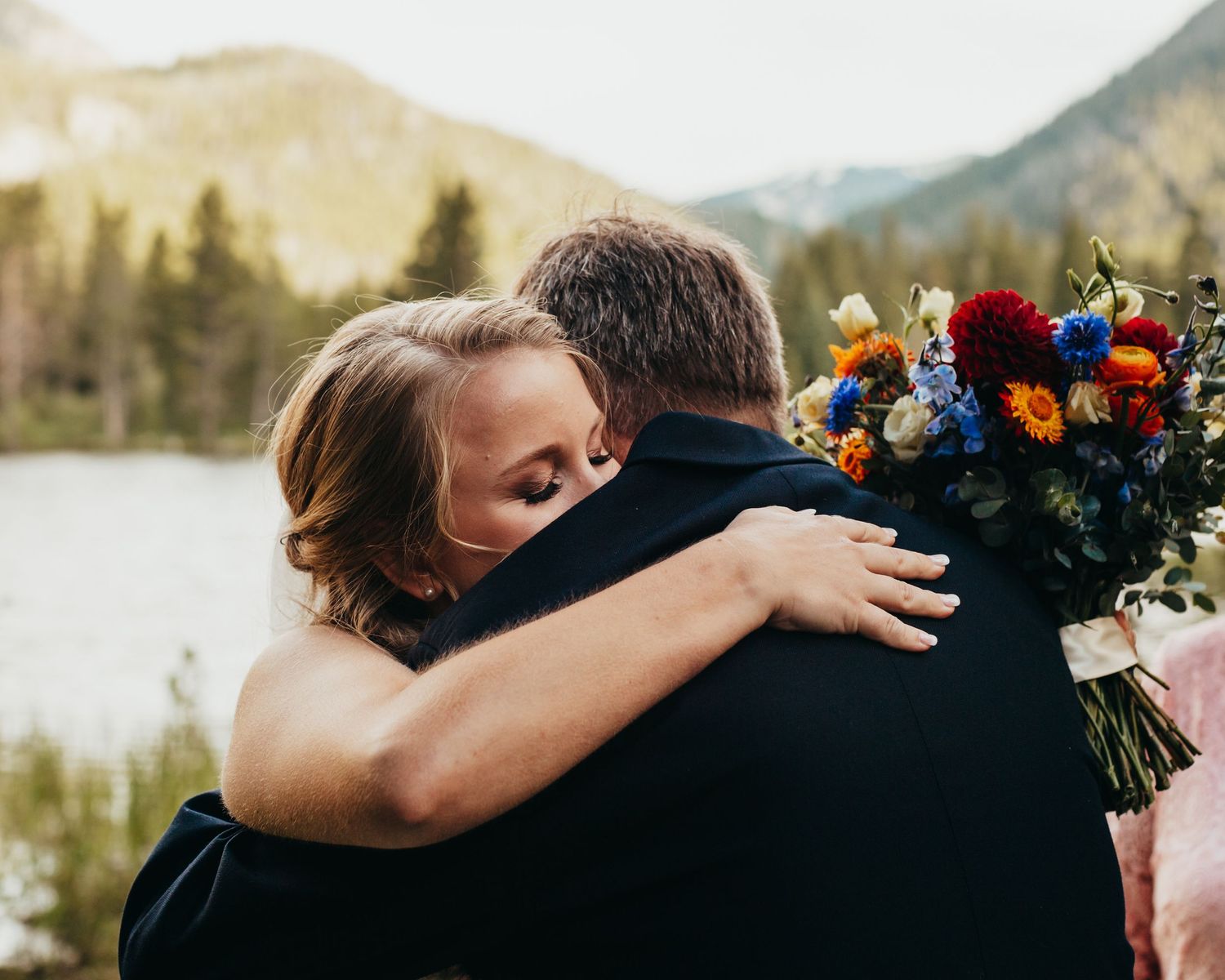 Intimate embrace with colorful wedding bouquet against mountain lake backdrop.