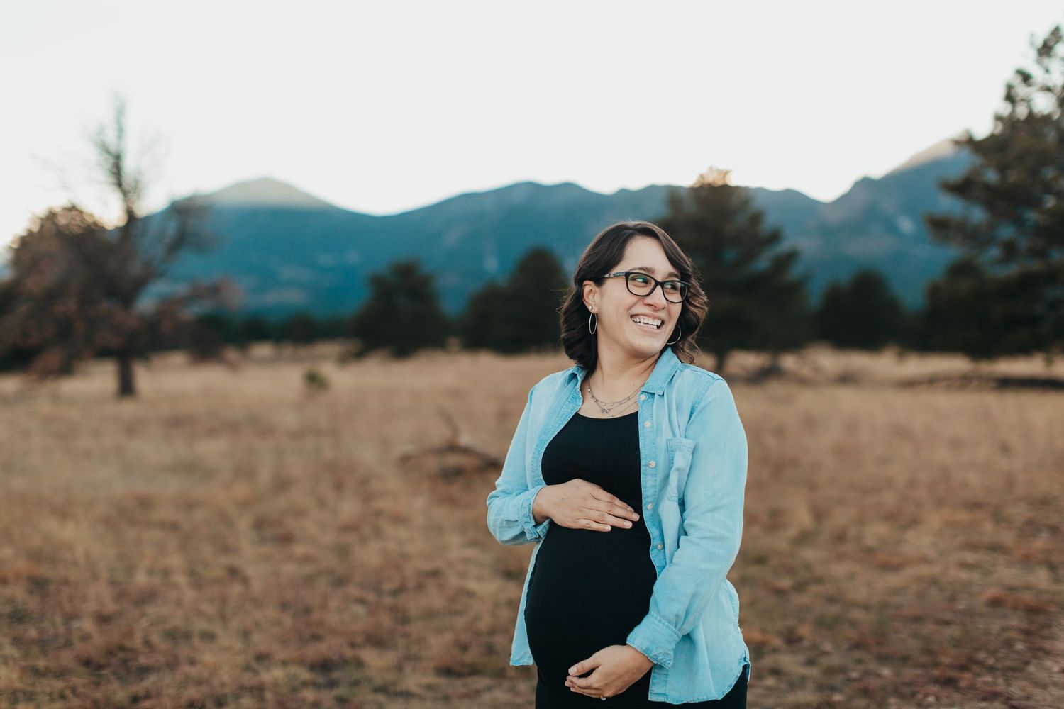 Expectant mother wearing glasses and blue cardigan poses in a field with mountains in background.