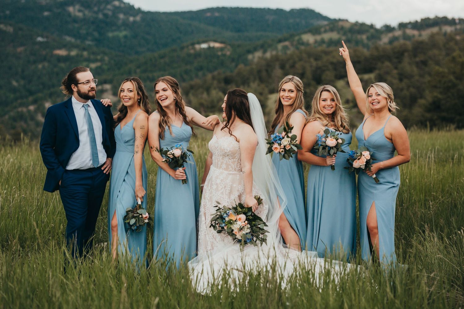 Wedding party in light blue dresses and navy suits poses together in a mountain meadow.