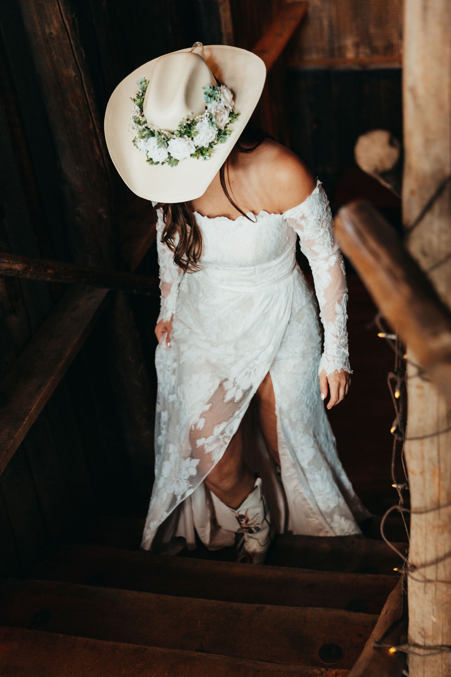 A lace wedding dress and white cowboy hat with floral accents on wooden stairs in rustic setting.