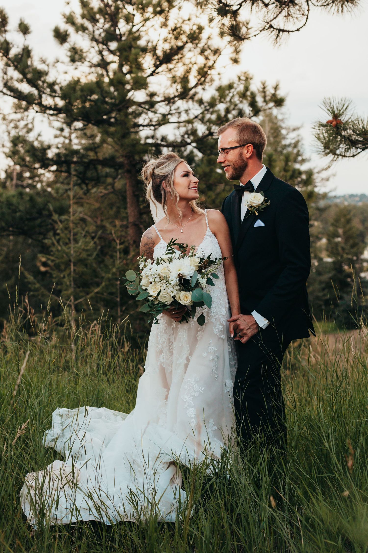 A newlywed couple poses together in tall grass with pine trees in the background.