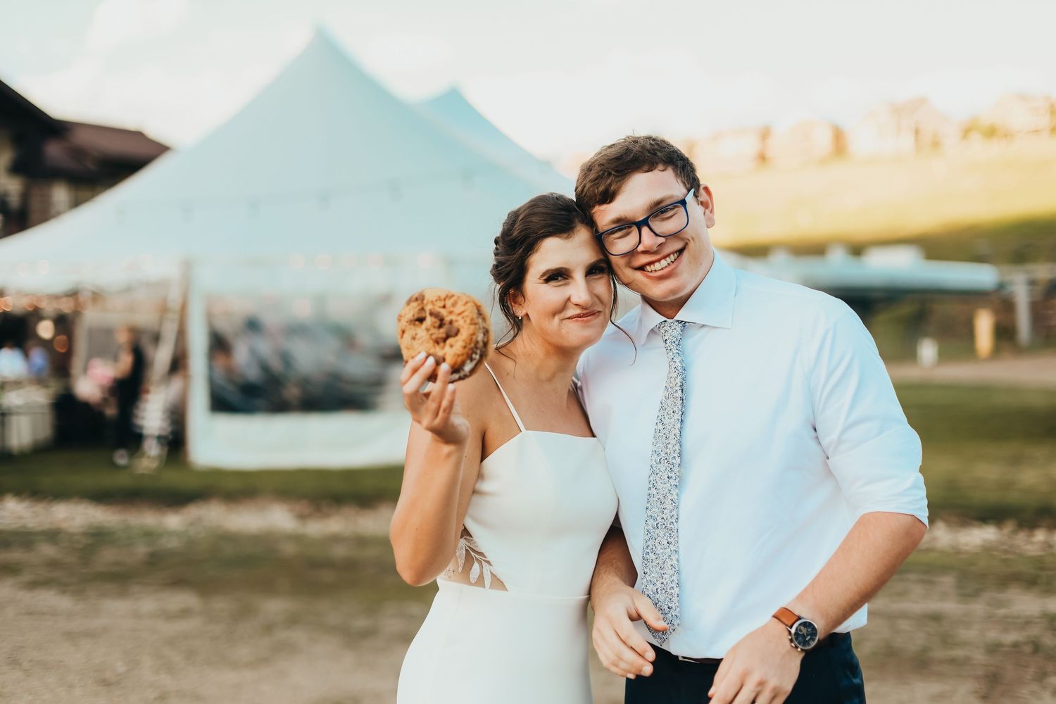 A couple at an outdoor wedding venue shares a burger while wearing formal attire.
