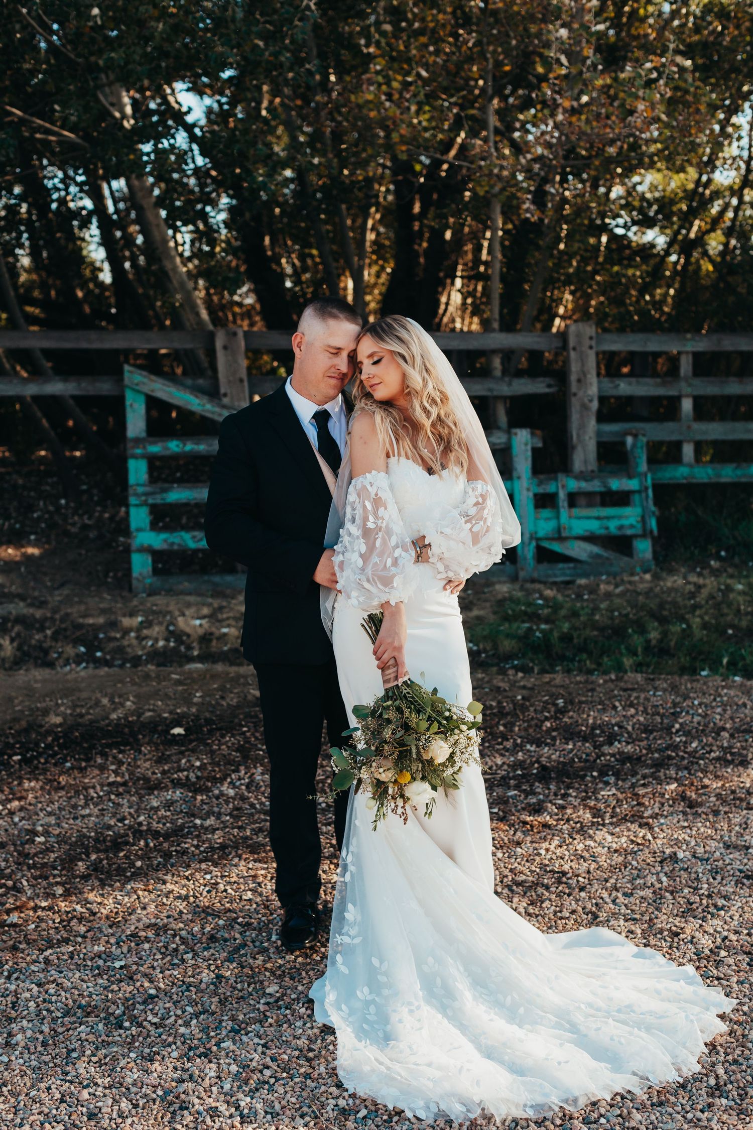Romantic wedding portrait against rustic fence backdrop in autumn forest setting.