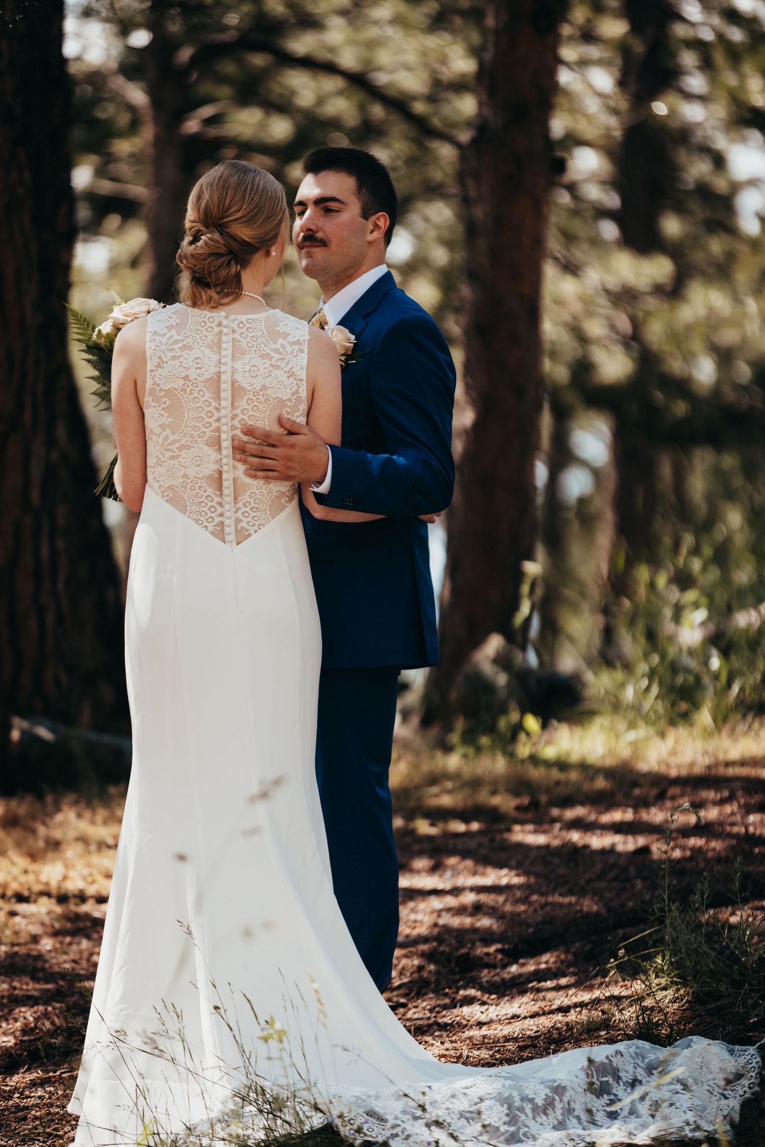 A couple shares a tender moment in a forest setting, with the bride wearing a lace-back wedding gown.