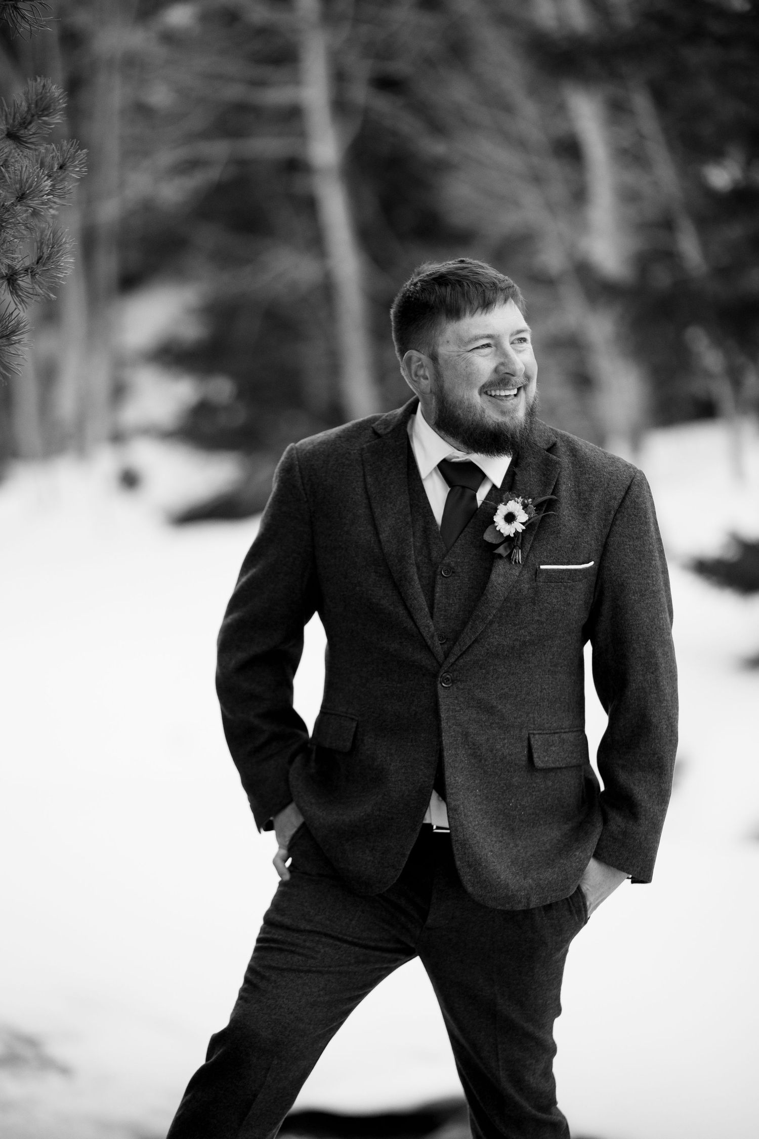 A groom in a dark suit poses against a snowy winter landscape background.