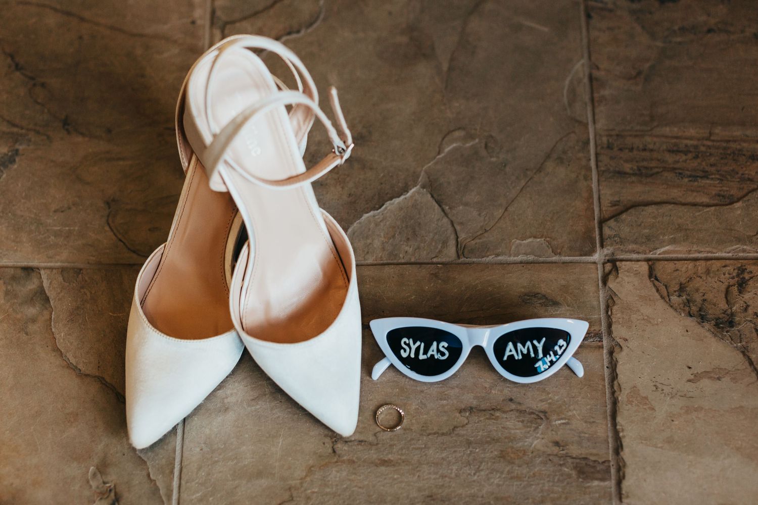 Elegant white bridal shoes and retro sunglasses displayed on stone tiles.