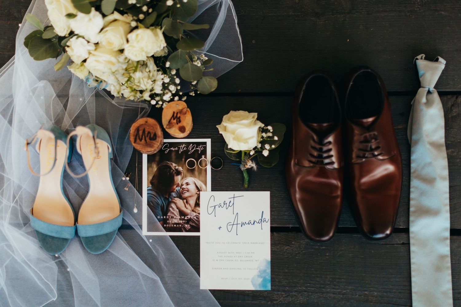 Wedding accessories including blue heels, brown dress shoes, veil and floral arrangements on dark surface.