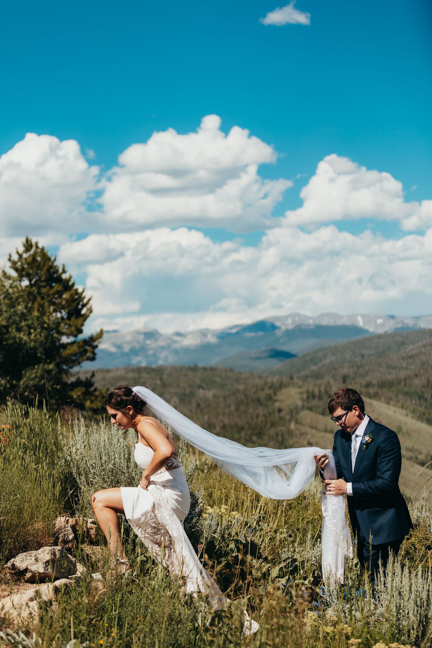 A couple poses on rocky mountain terrain with a flowing veil against blue skies.