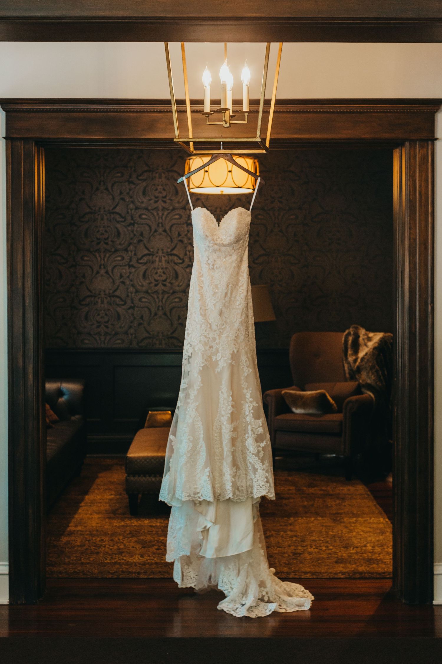 Elegant white lace wedding dress hanging beneath chandelier in dramatic lighting.