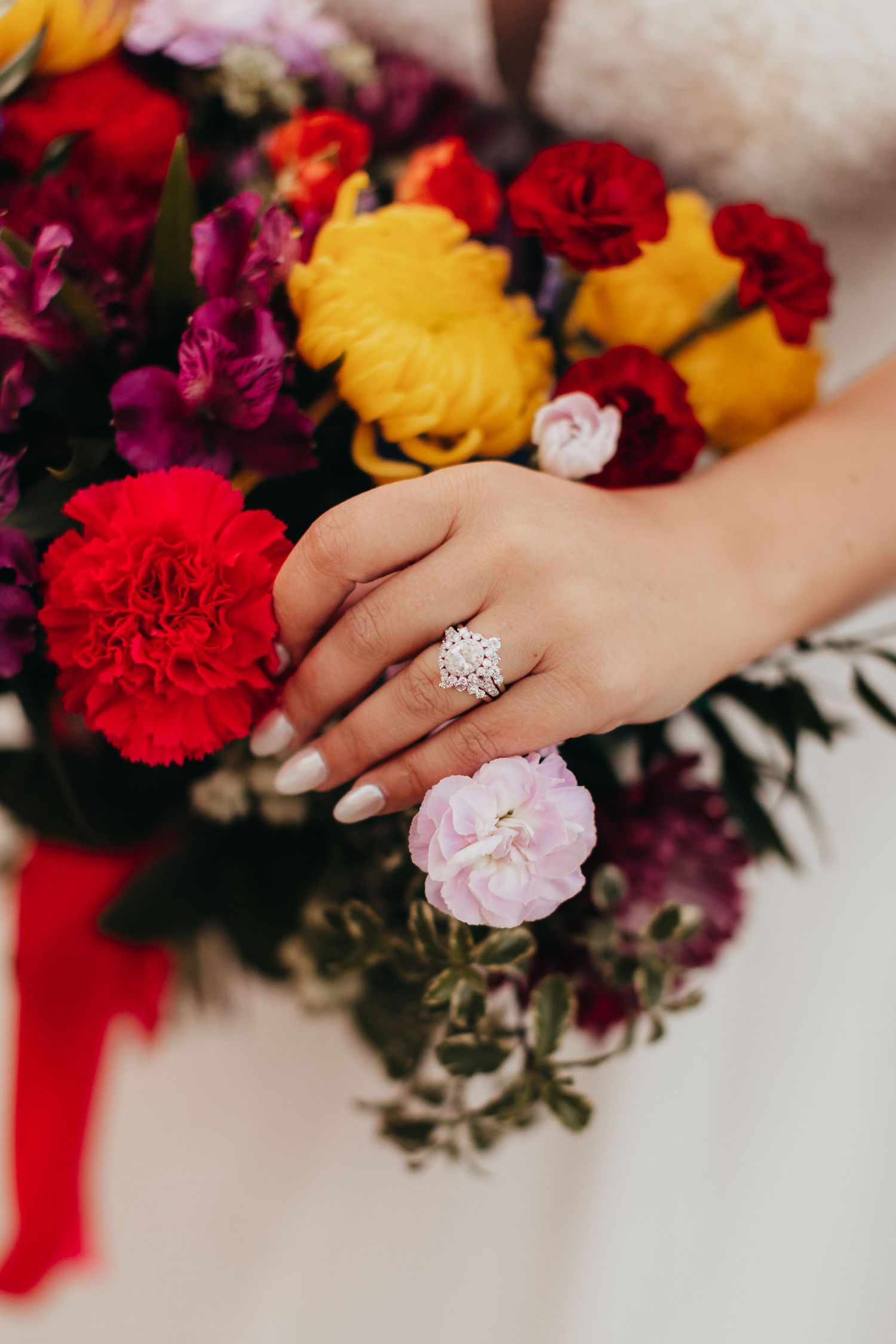 Close-up of a hand wearing a diamond engagement ring holding a vibrant bouquet with red and yellow flowers.