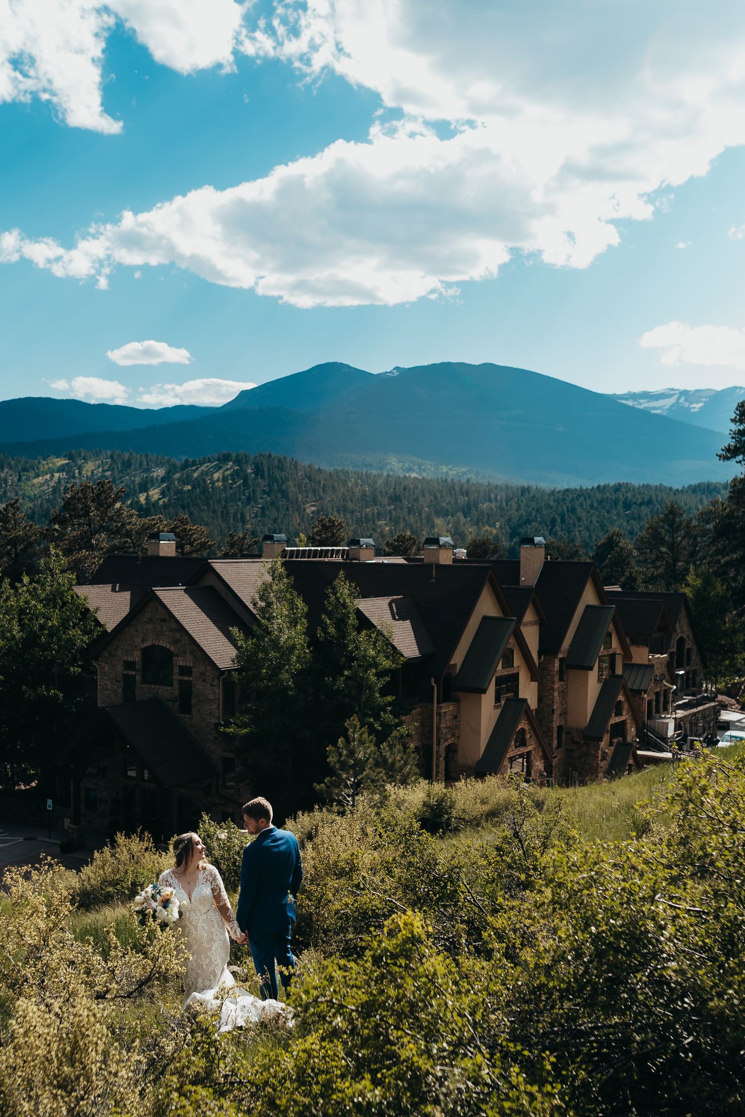 A couple descends a mountain path with rustic lodges and blue skies in the background.