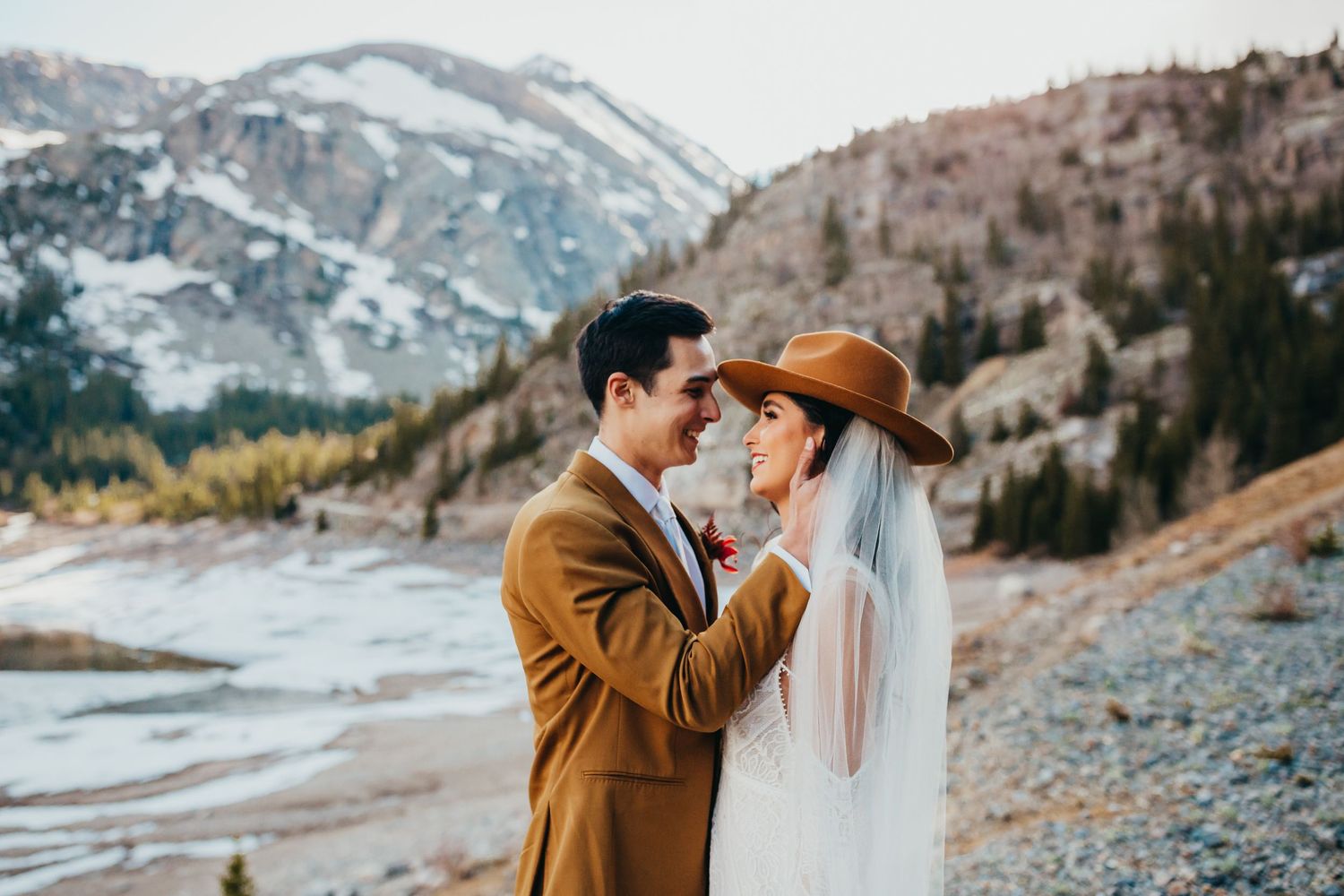 A romantic wedding moment captured in front of snow-capped mountains with a brown suit and white dress.