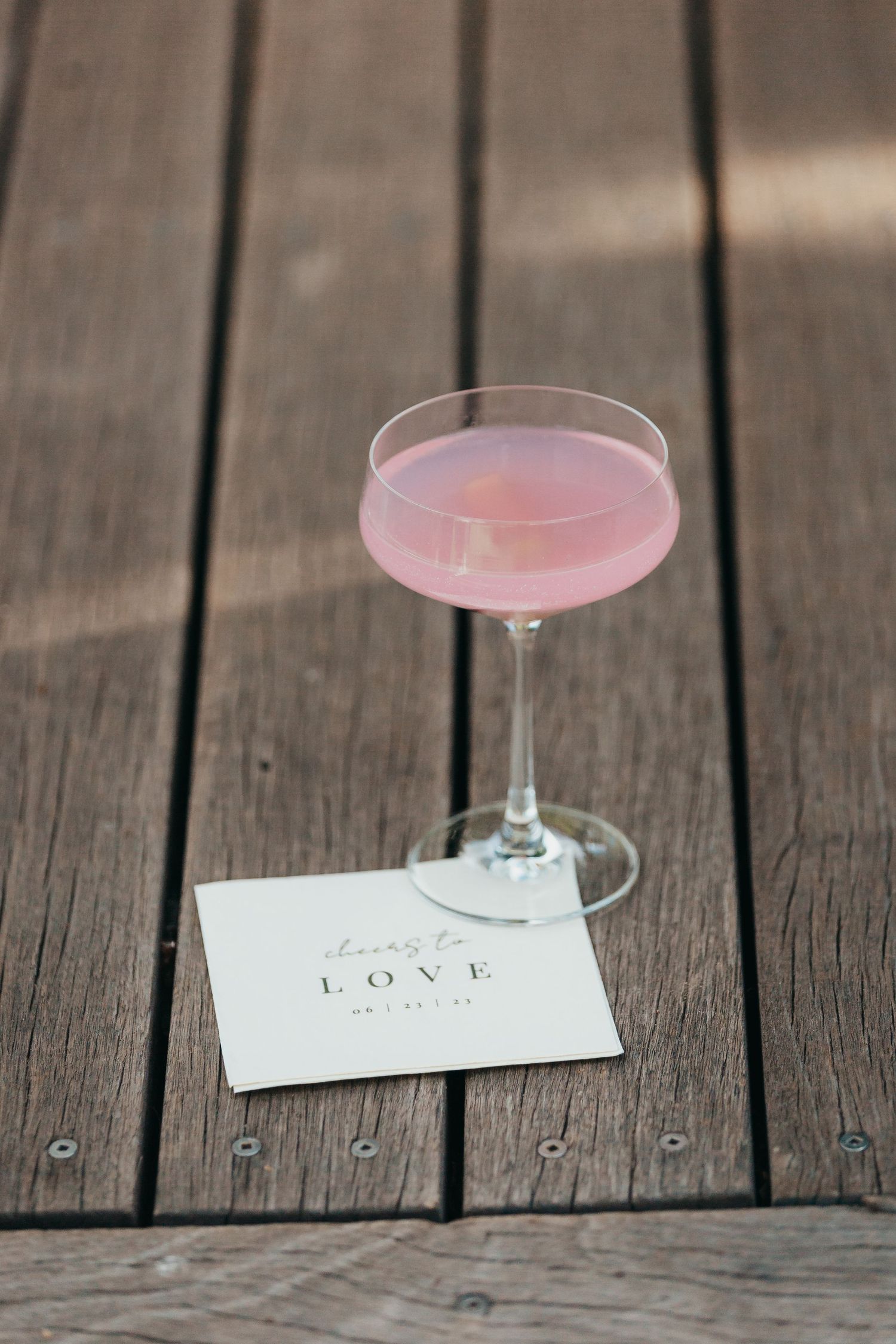 Pink cocktail and love note on wooden deck at wedding reception.