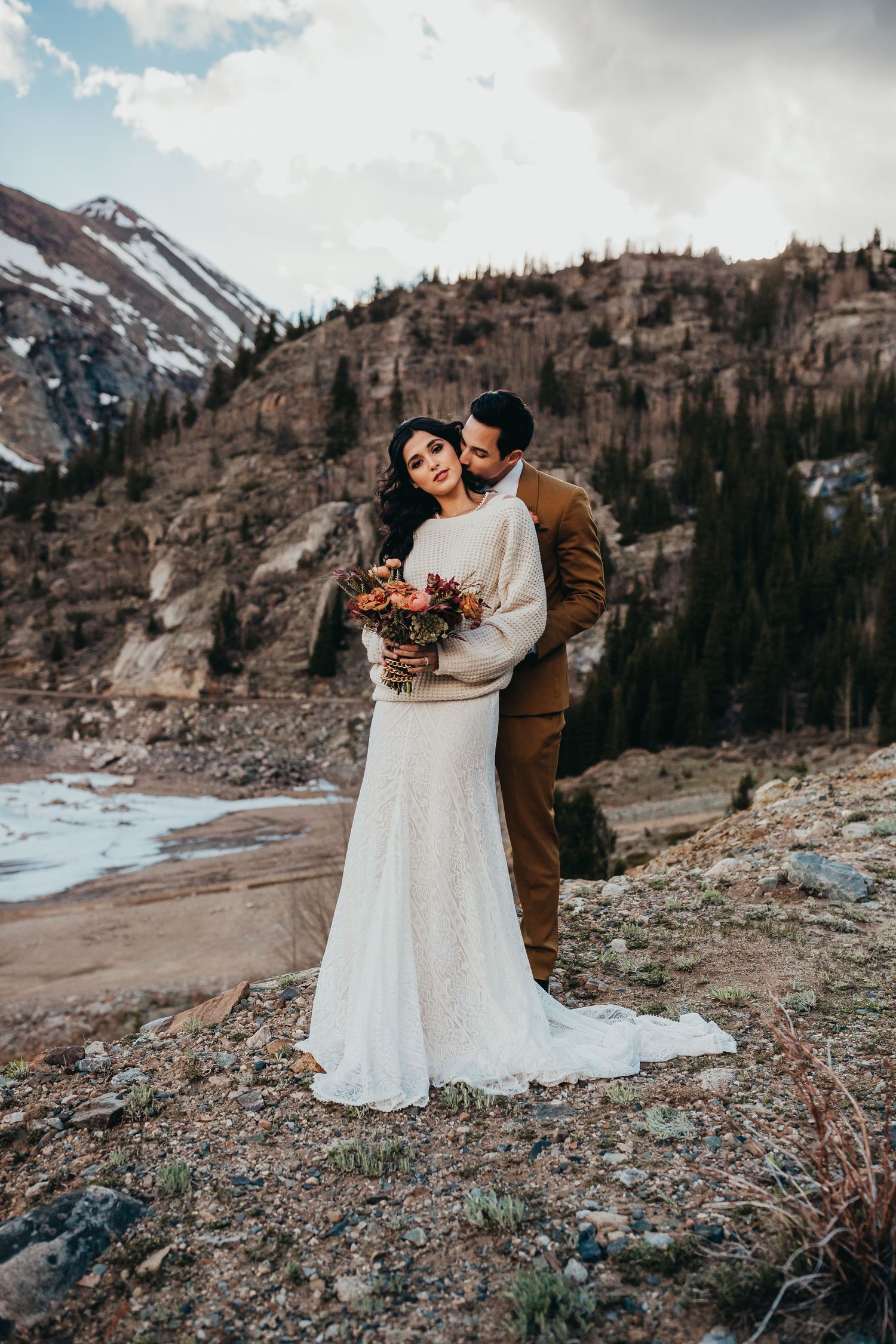 Wedding couple embraces on rocky mountain terrain with autumn bouquet and dramatic landscape.