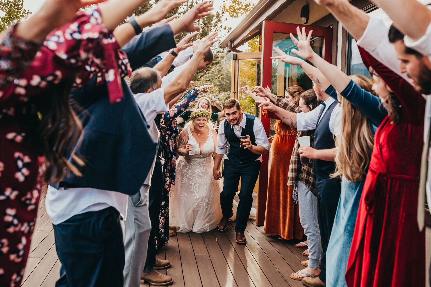 Wedding guests form a celebratory tunnel with raised arms on a wooden deck.