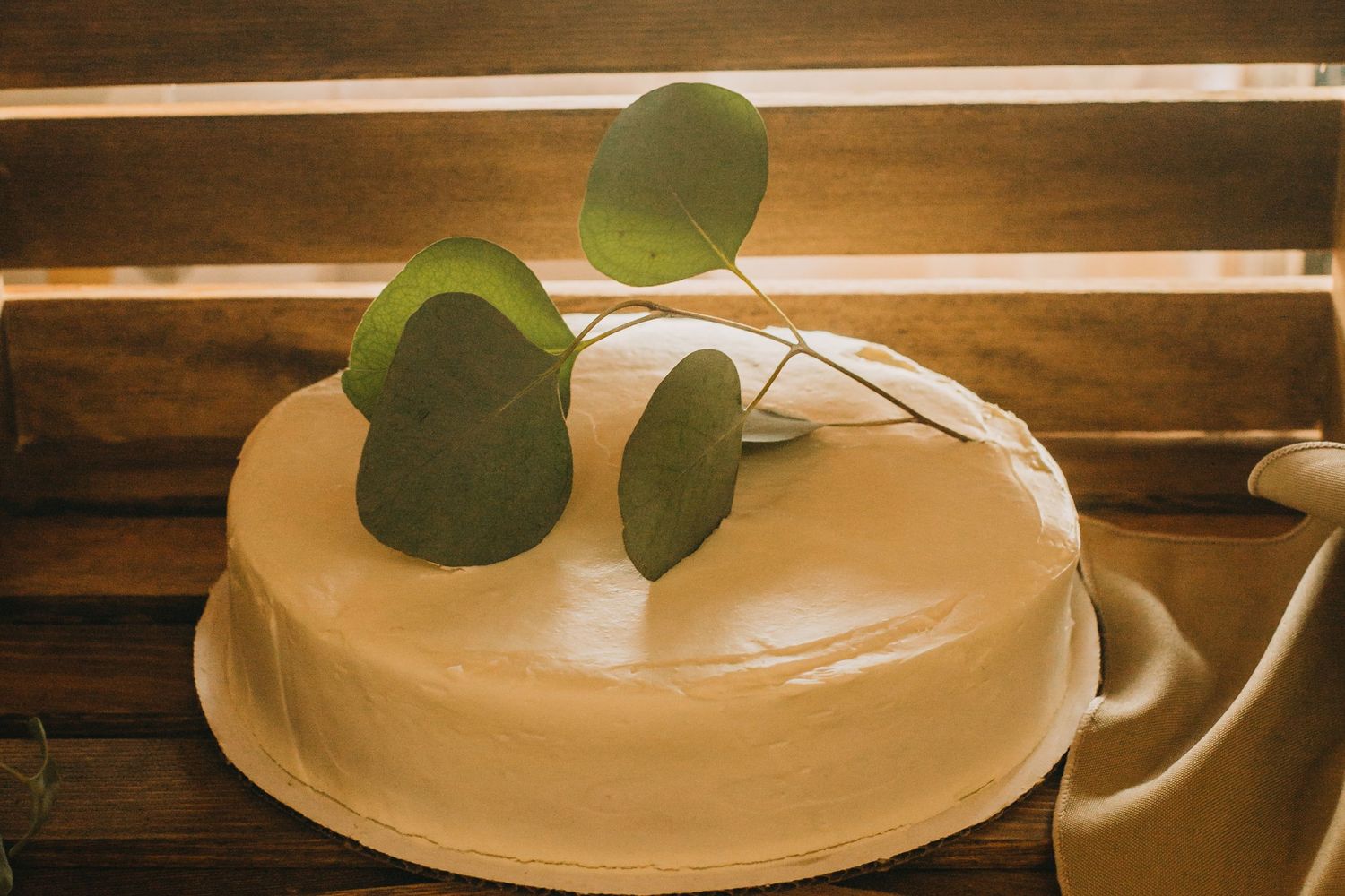 A simple white frosted cake decorated with fresh eucalyptus leaves on a wooden surface.