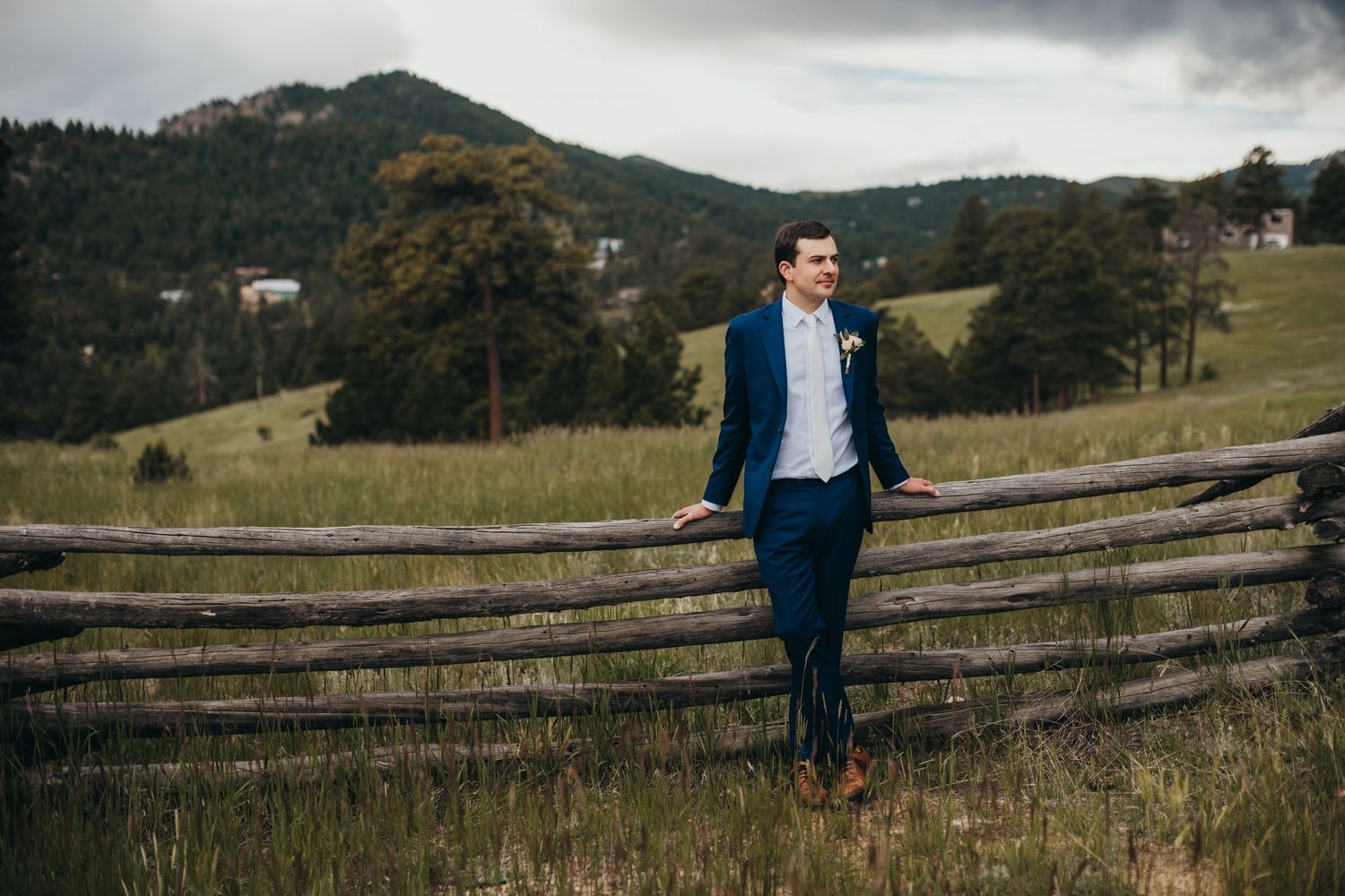 Groom leaning on rustic wooden fence with mountain vista in the background.