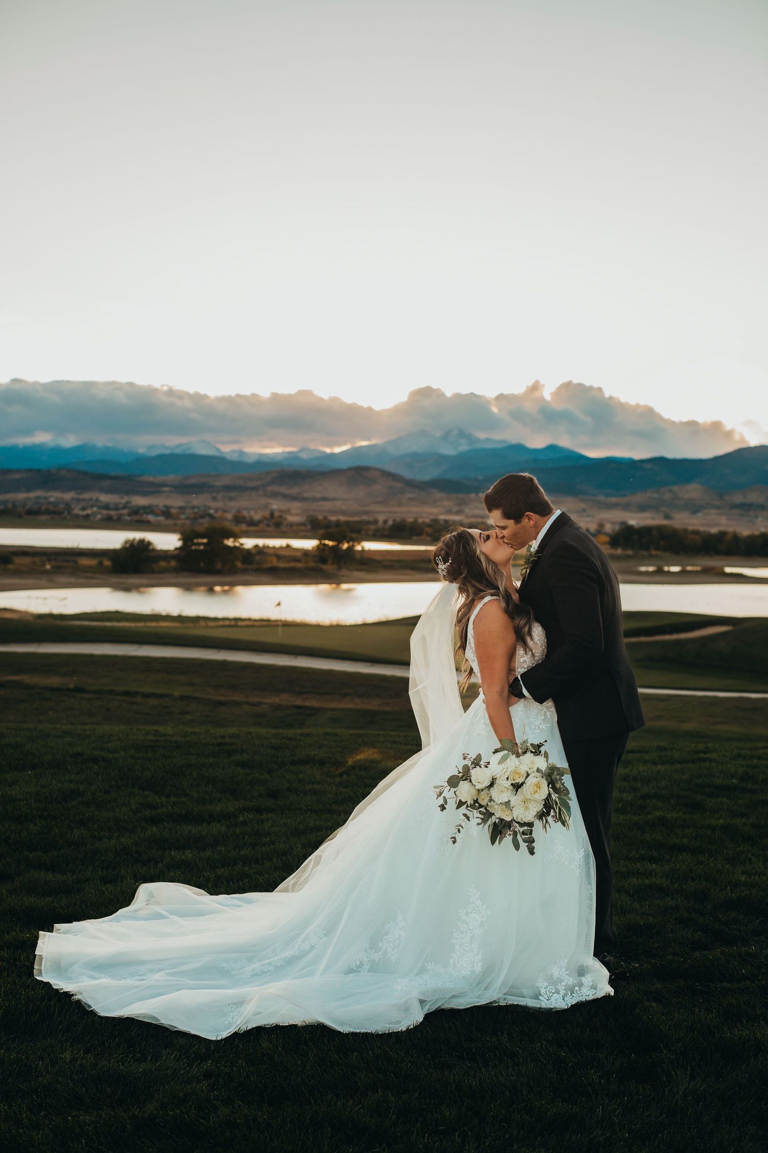 A wedding couple shares an intimate moment at sunset with mountains and a lake in the distance.