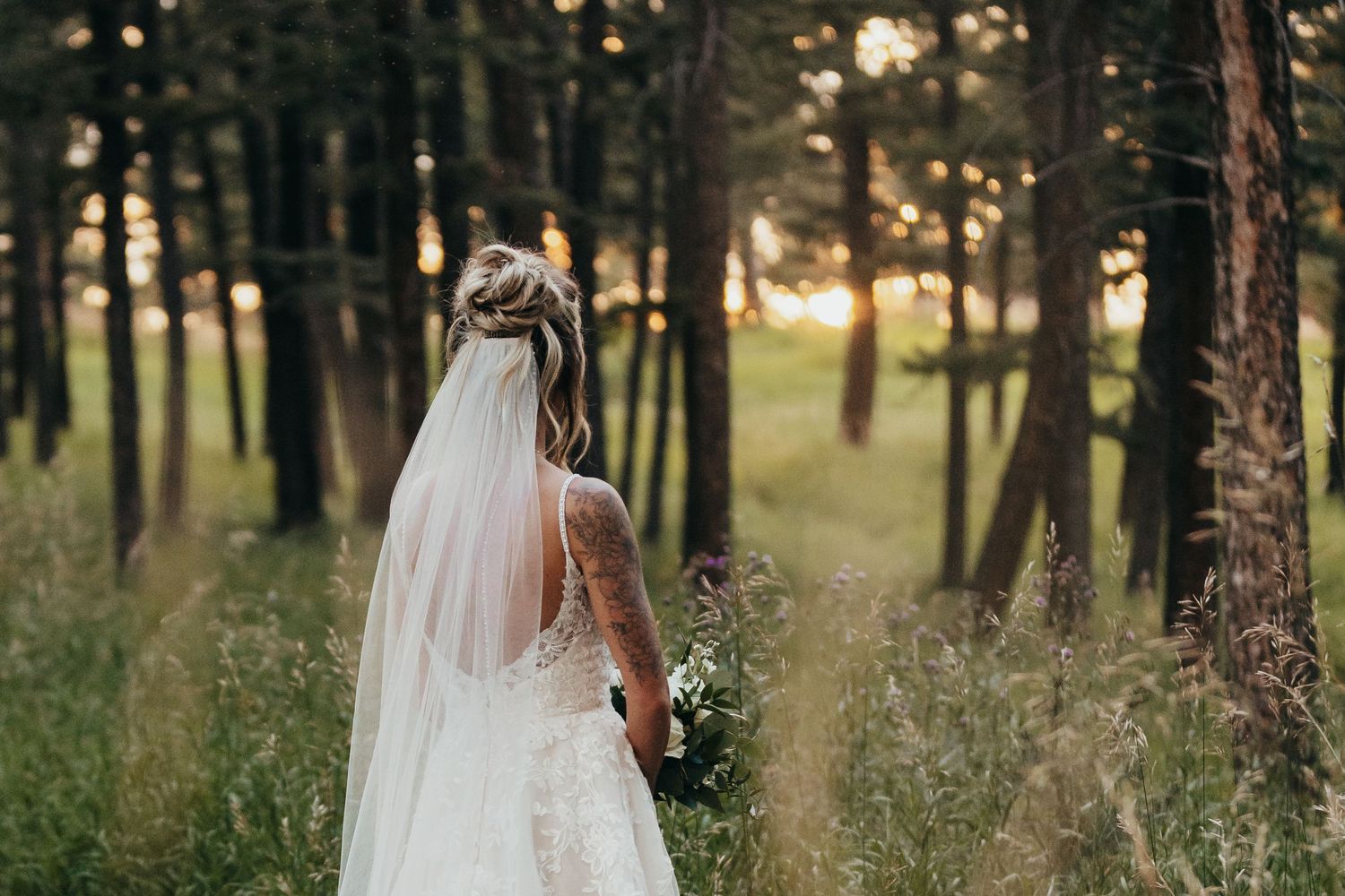 A bride in a white lace dress and veil walks through a sunlit forest.