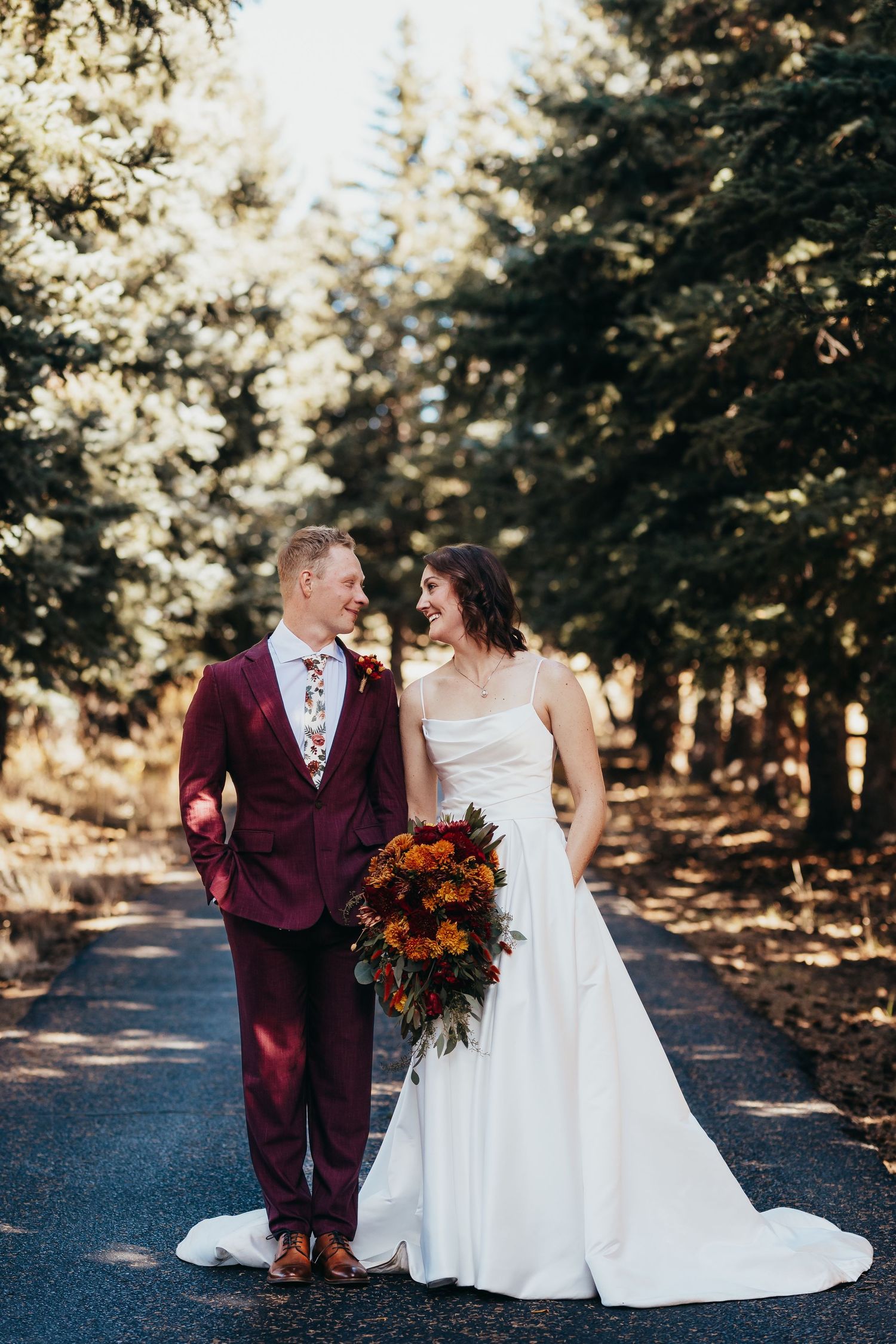 Couple in burgundy suit and white dress holding autumn-colored bouquet on forest path.