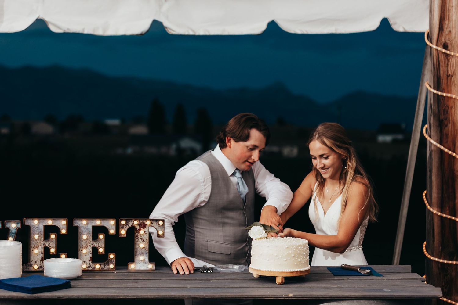 Couple shares intimate moment while cutting wedding cake under evening tent lighting.