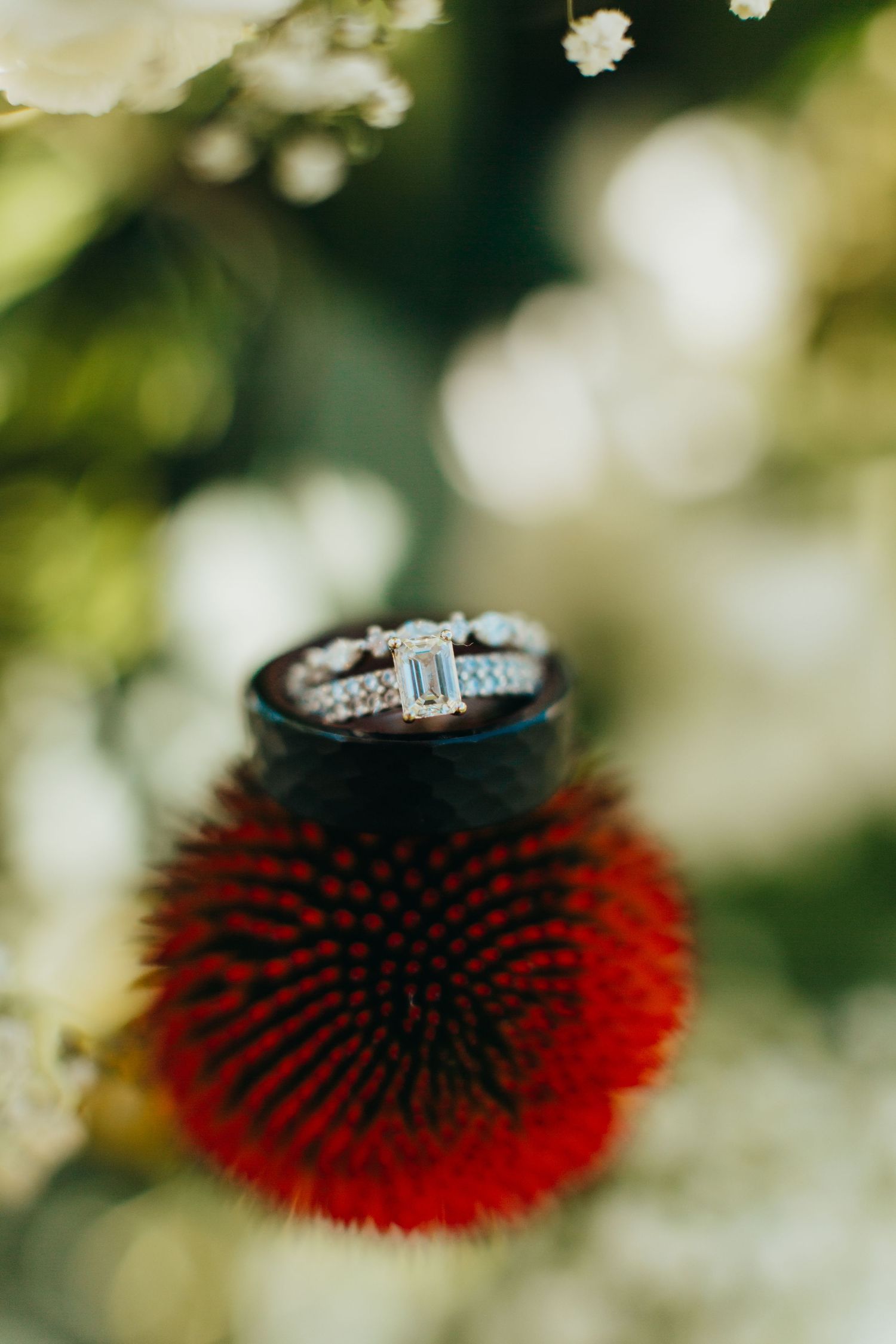 Wedding ring rests atop a vibrant red echinacea flower with blurred bokeh background.