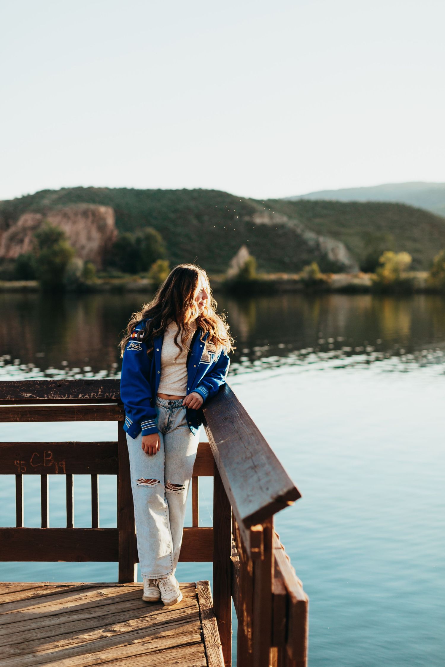 Person in blue jacket standing on wooden dock overlooking serene lake and mountain landscape.