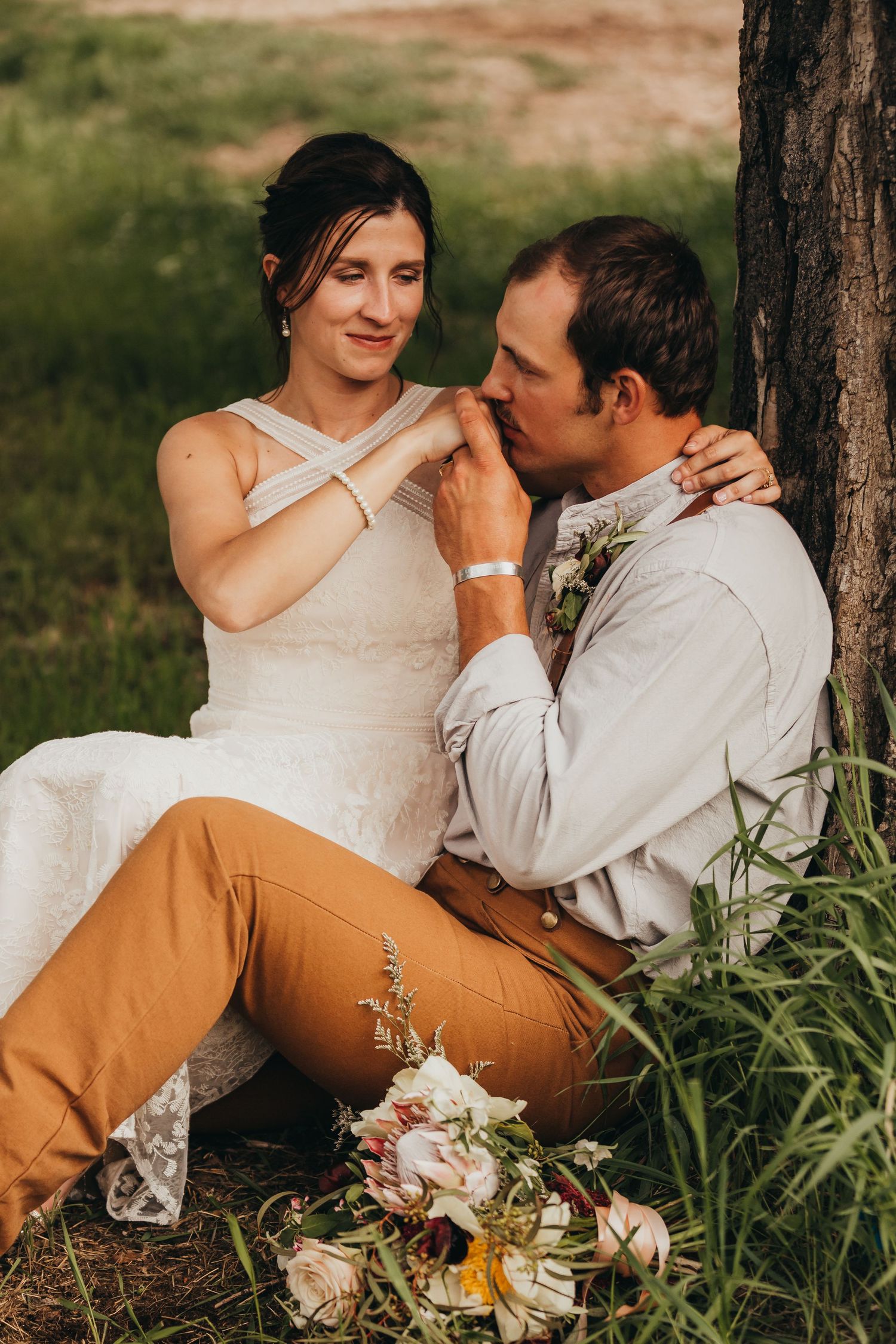 A romantic moment captured between a couple sitting under a tree with a floral bouquet nearby.