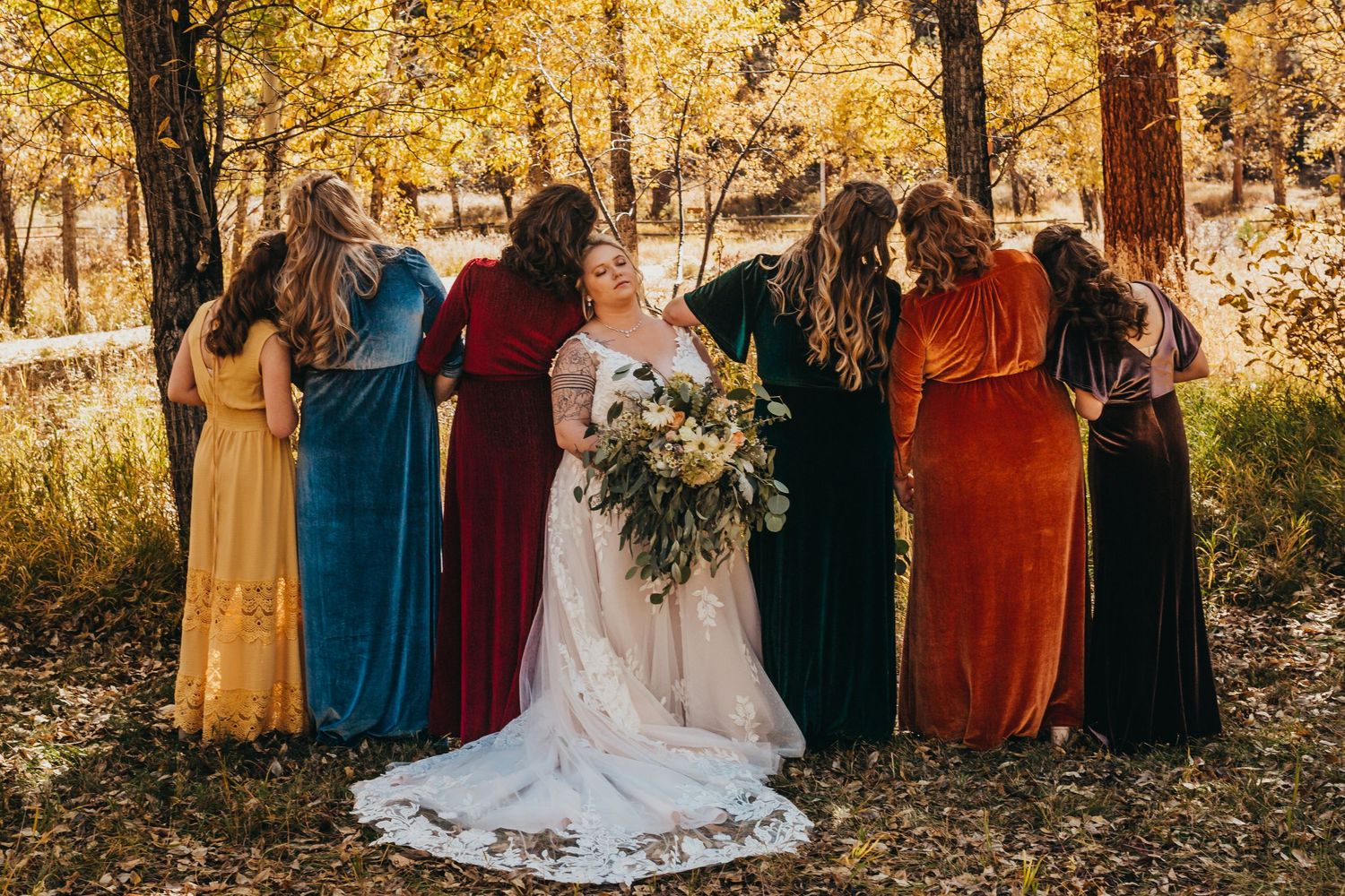 Wedding party poses in rich, jewel-toned dresses against a backdrop of golden autumn trees.