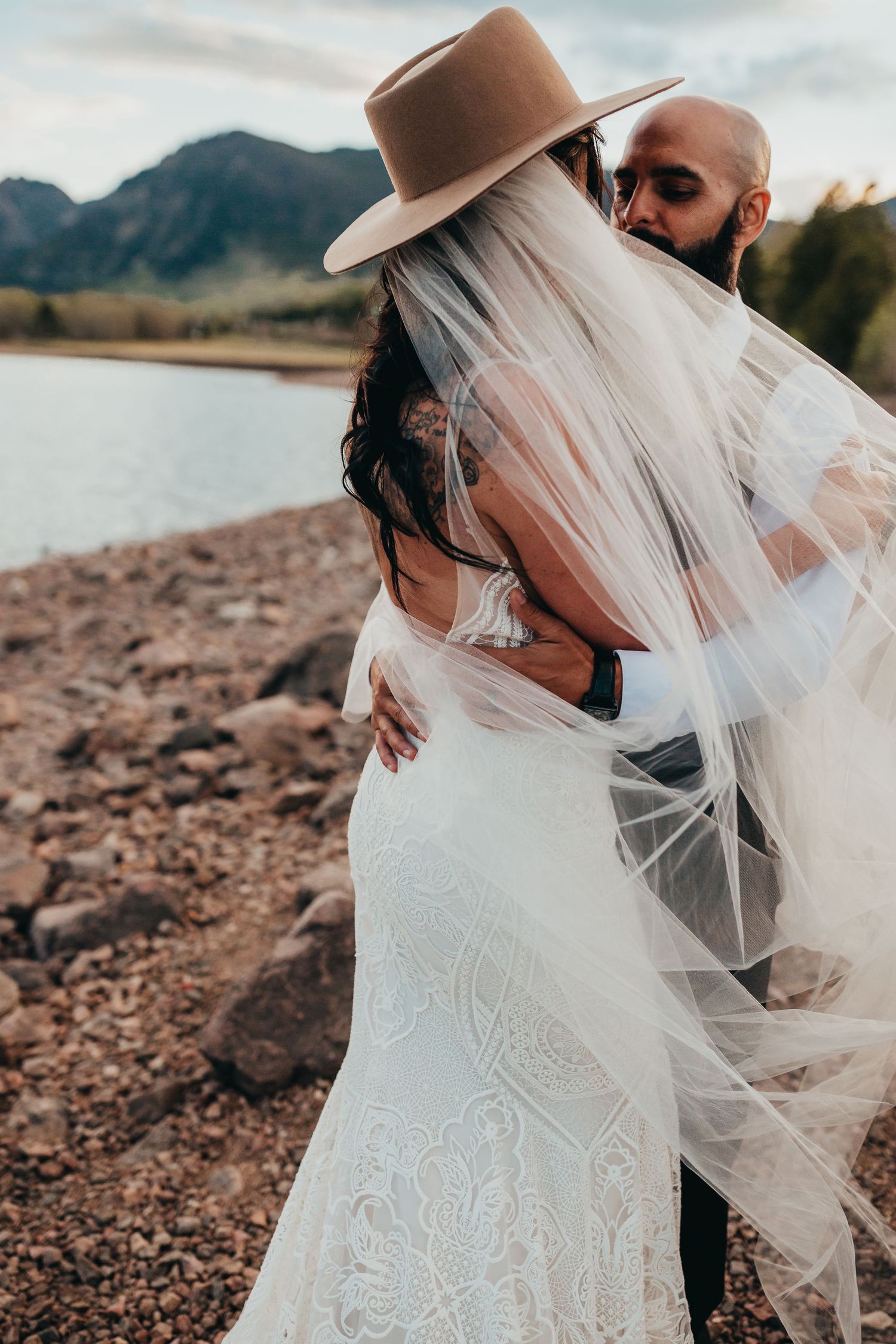Intimate wedding moment with flowing veil and brown hat against mountain lake backdrop.