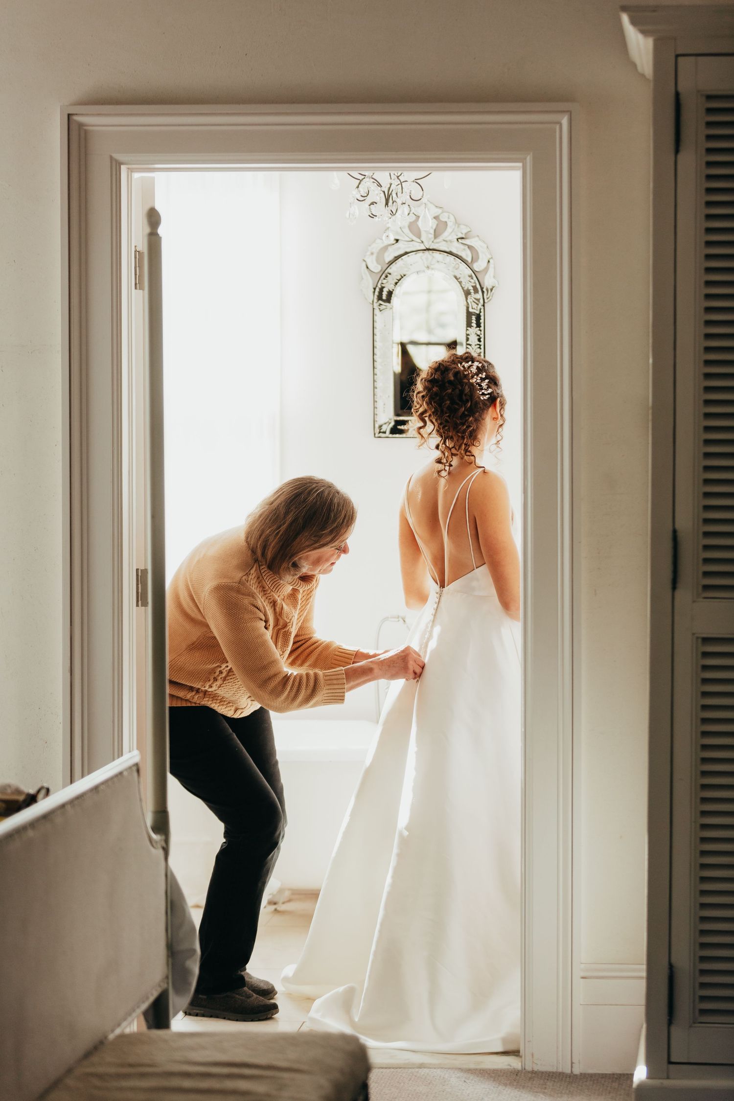Helper adjusts wedding dress train in sunlit doorway.
