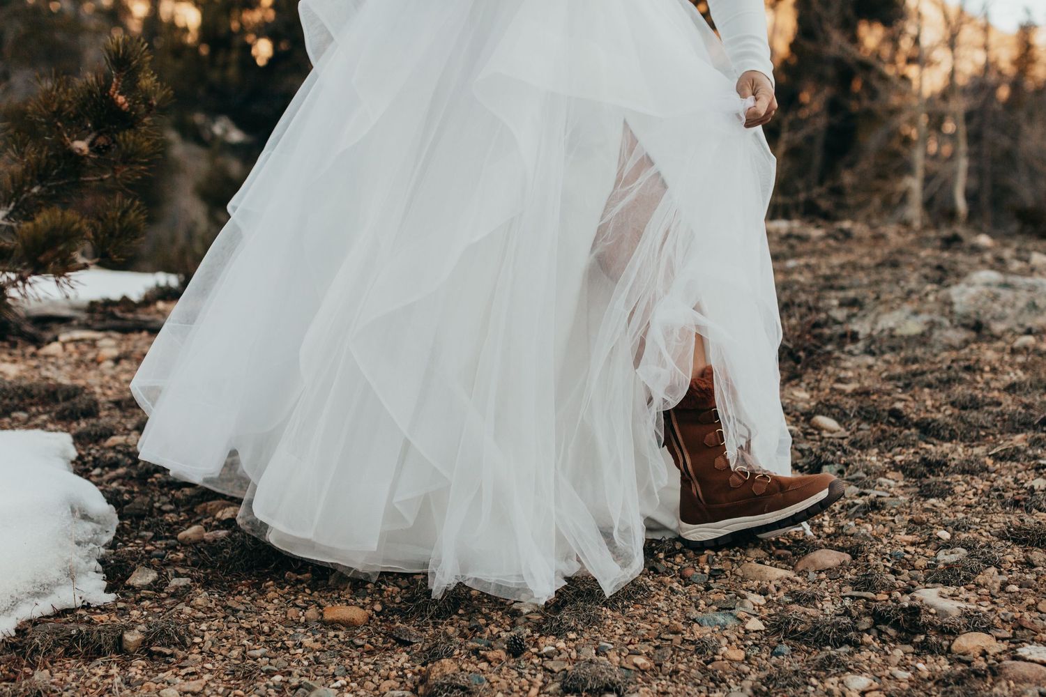 A white tulle wedding dress hem paired with brown leather boots in snowy winter ground.