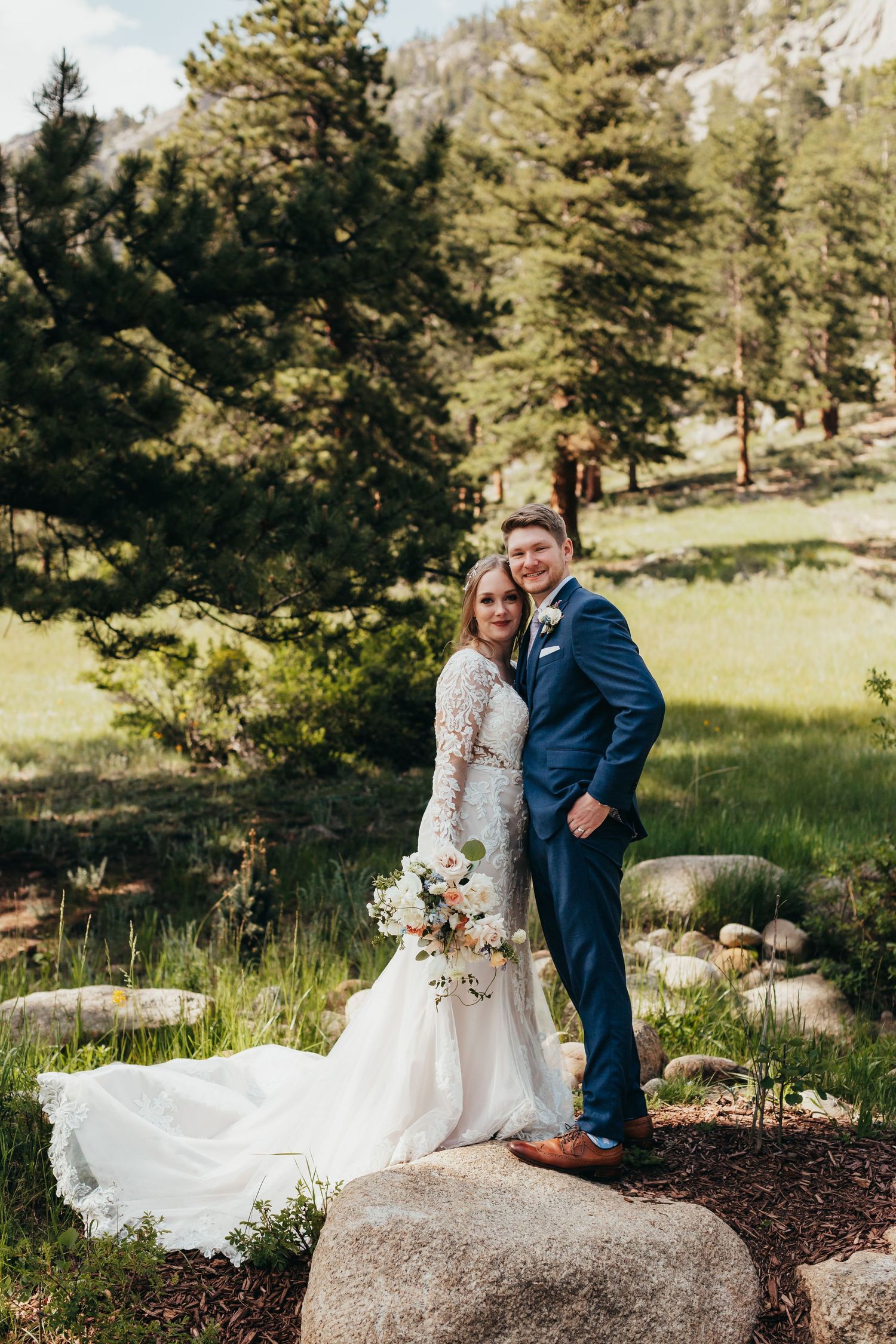 Couple in wedding attire poses on large boulder in sunlit mountain meadow surrounded by pine trees.