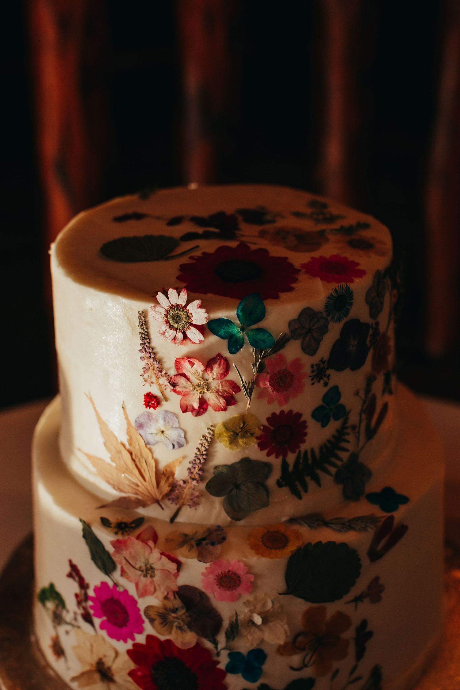 Two-tiered white wedding cake decorated with pressed flowers in vibrant colors.