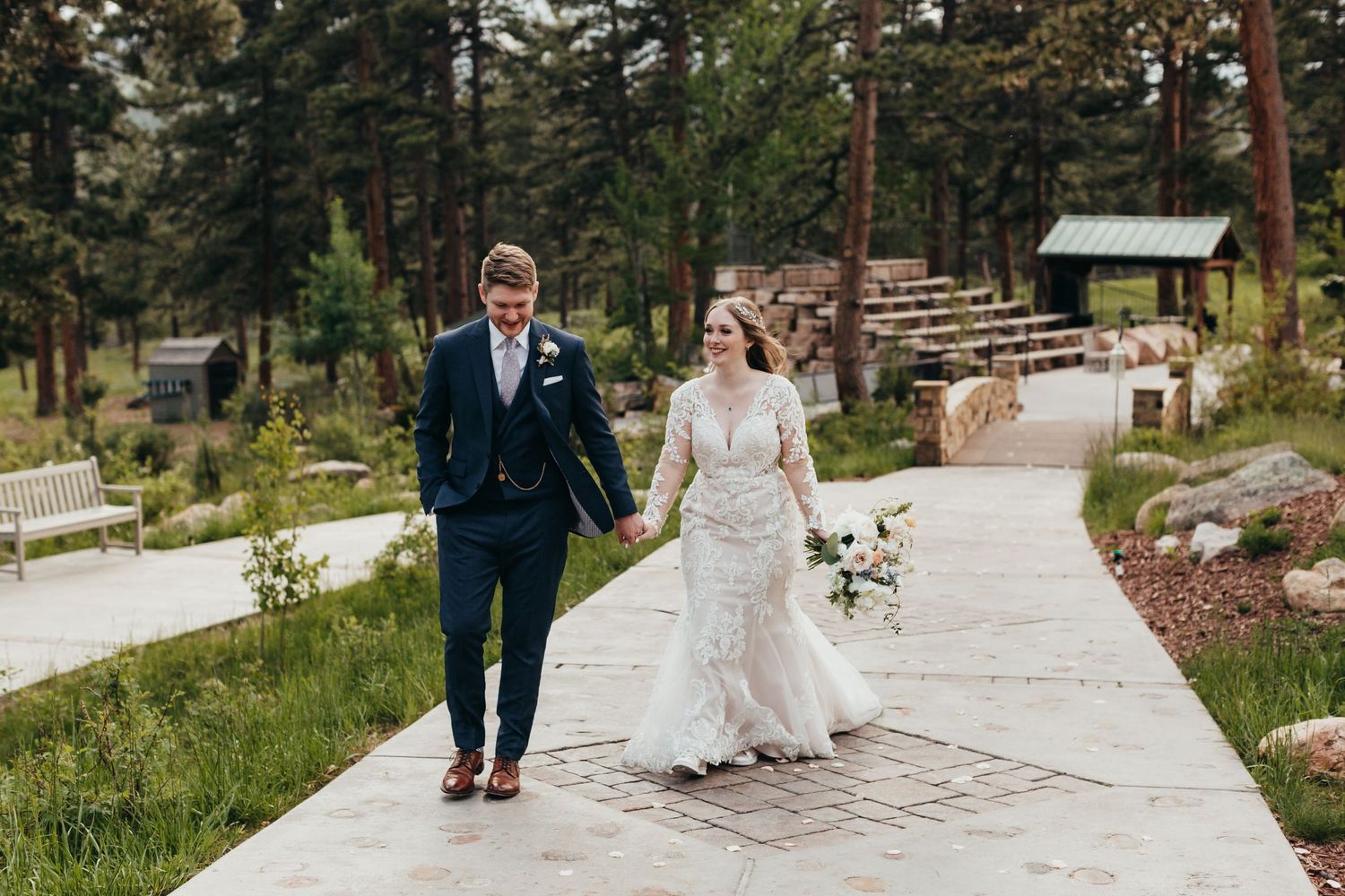 Newlyweds walking hand in hand on path surrounded by pine trees.