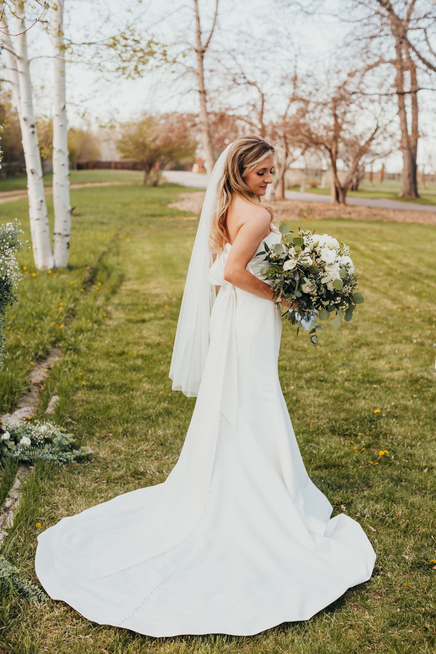 Beautiful bride in flowing white dress holding a lush bouquet in a garden with birch trees.