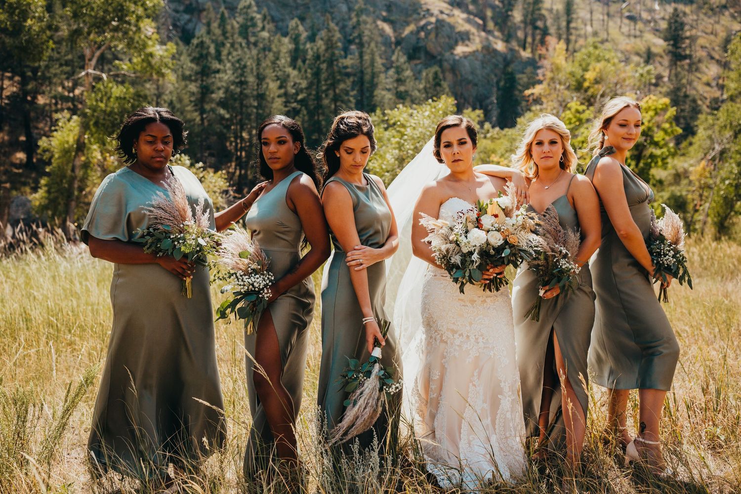 Bride and bridesmaids in neutral dresses gather in mountain meadow holding floral bouquets.