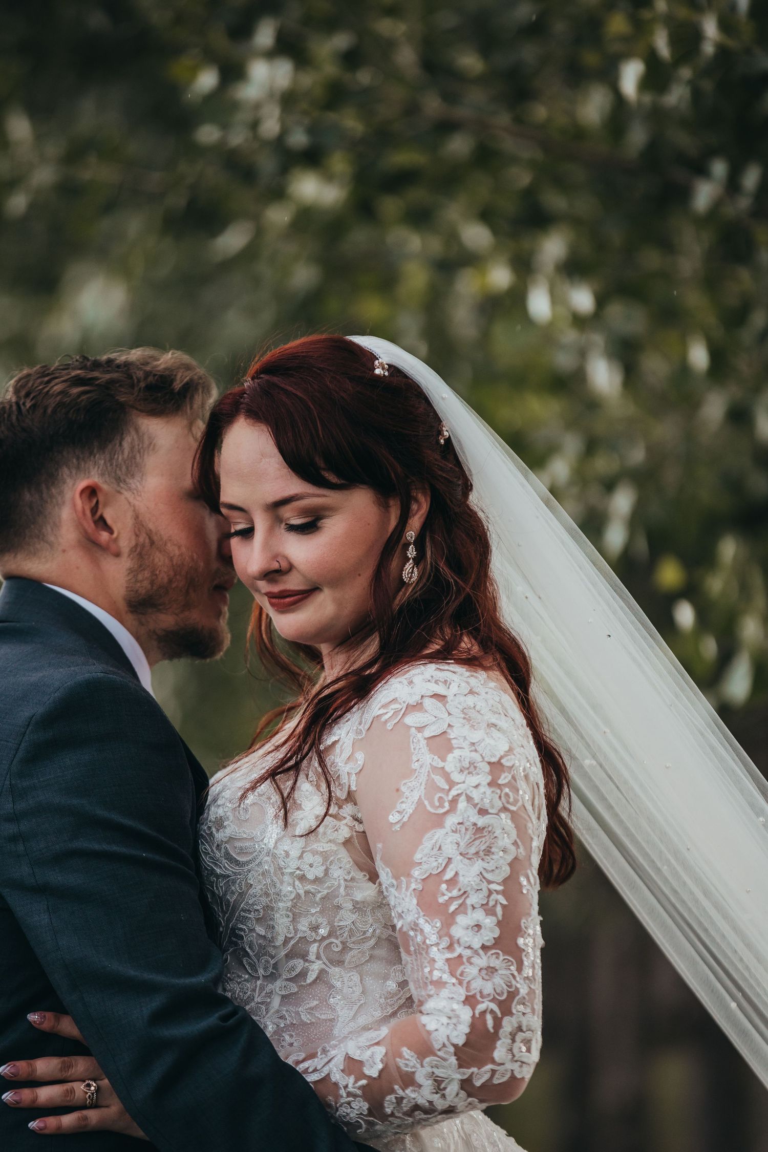 Intimate moment captured between couple in lace wedding dress and navy suit outdoors.