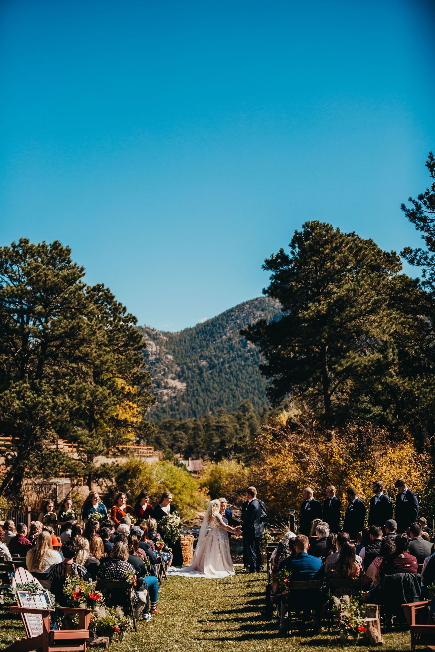 Outdoor mountain wedding ceremony with guests seated on wooden chairs under a bright blue sky.