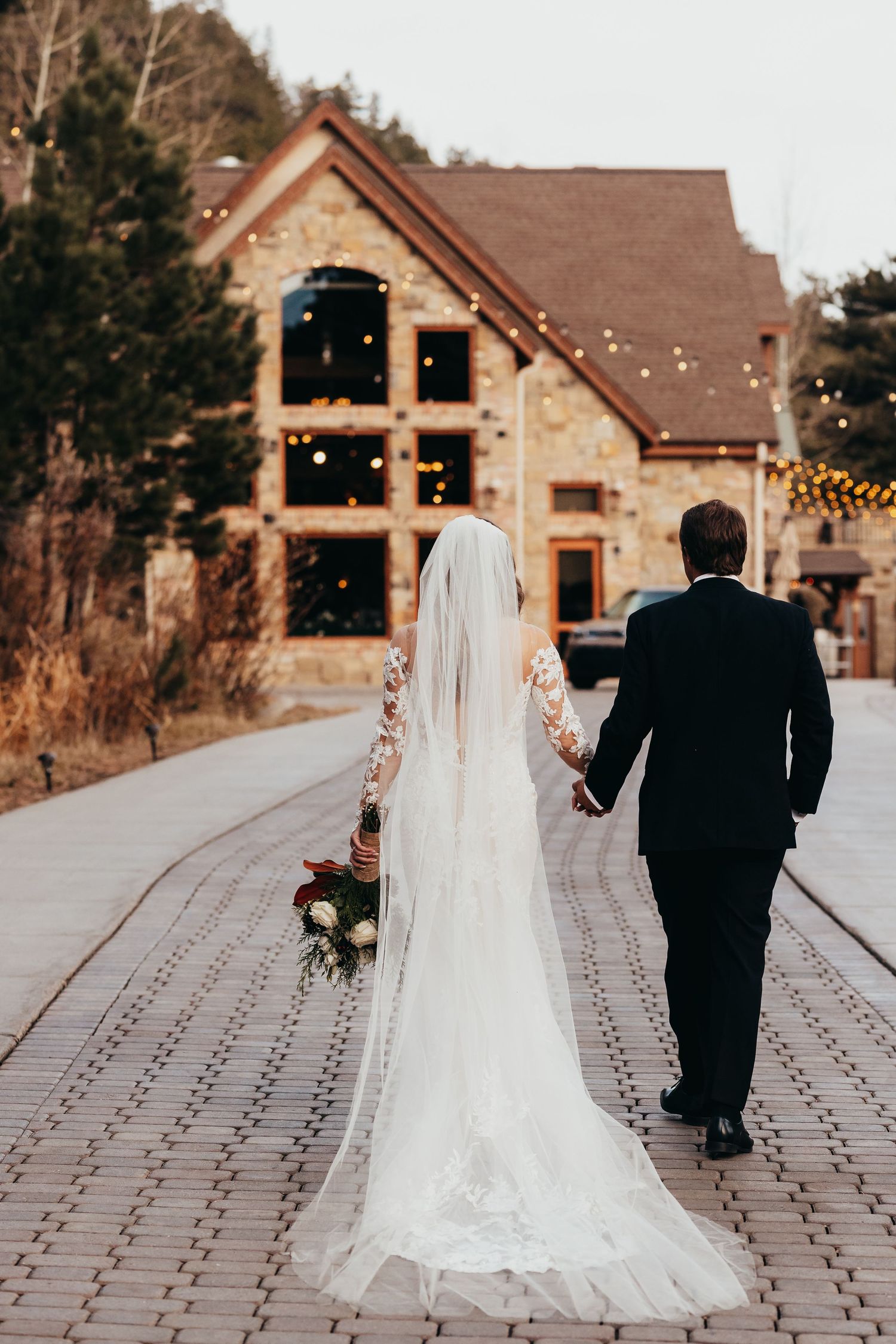 Newlyweds walk hand in hand towards rustic stone venue with string lights.