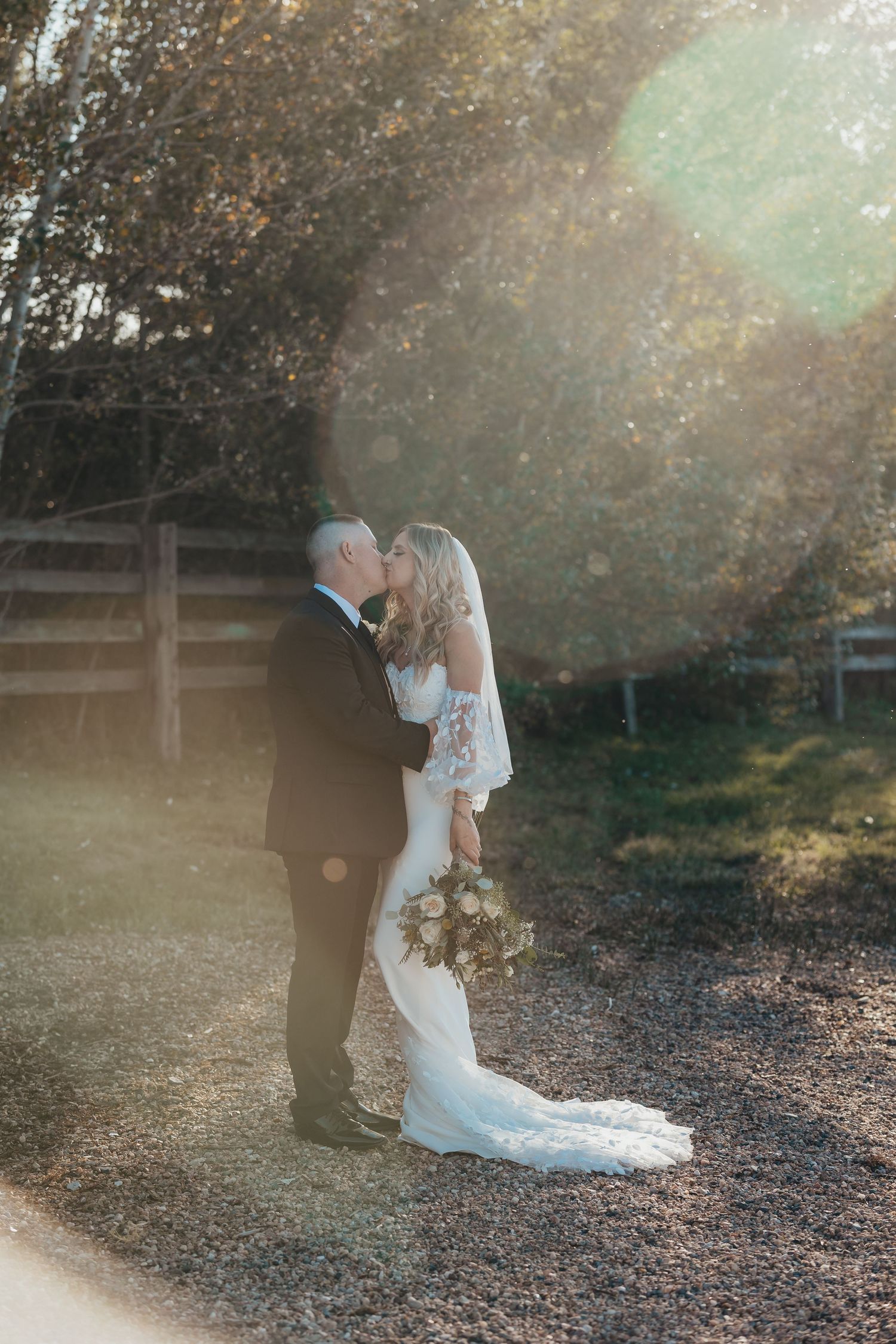 Sunlit wedding portrait with couple embracing on gravel path with lens flare effect.