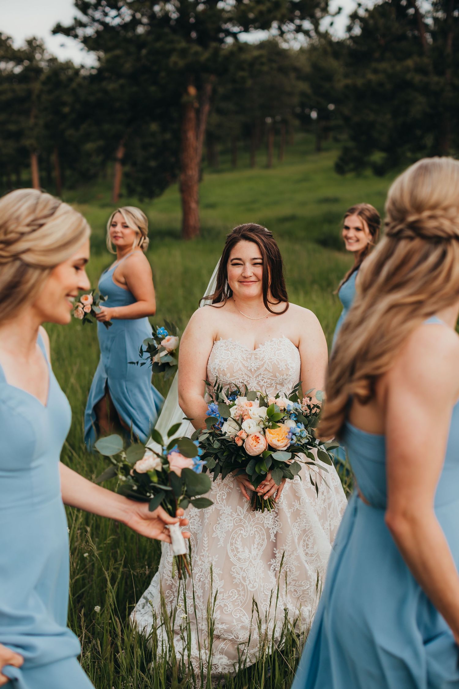 Wedding party in powder blue dresses gathered around bride holding peach and white bouquet in grassy field.