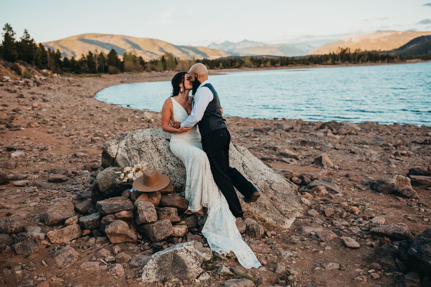 Newlyweds embrace on rocky lakeside with mountains and sunset in background.