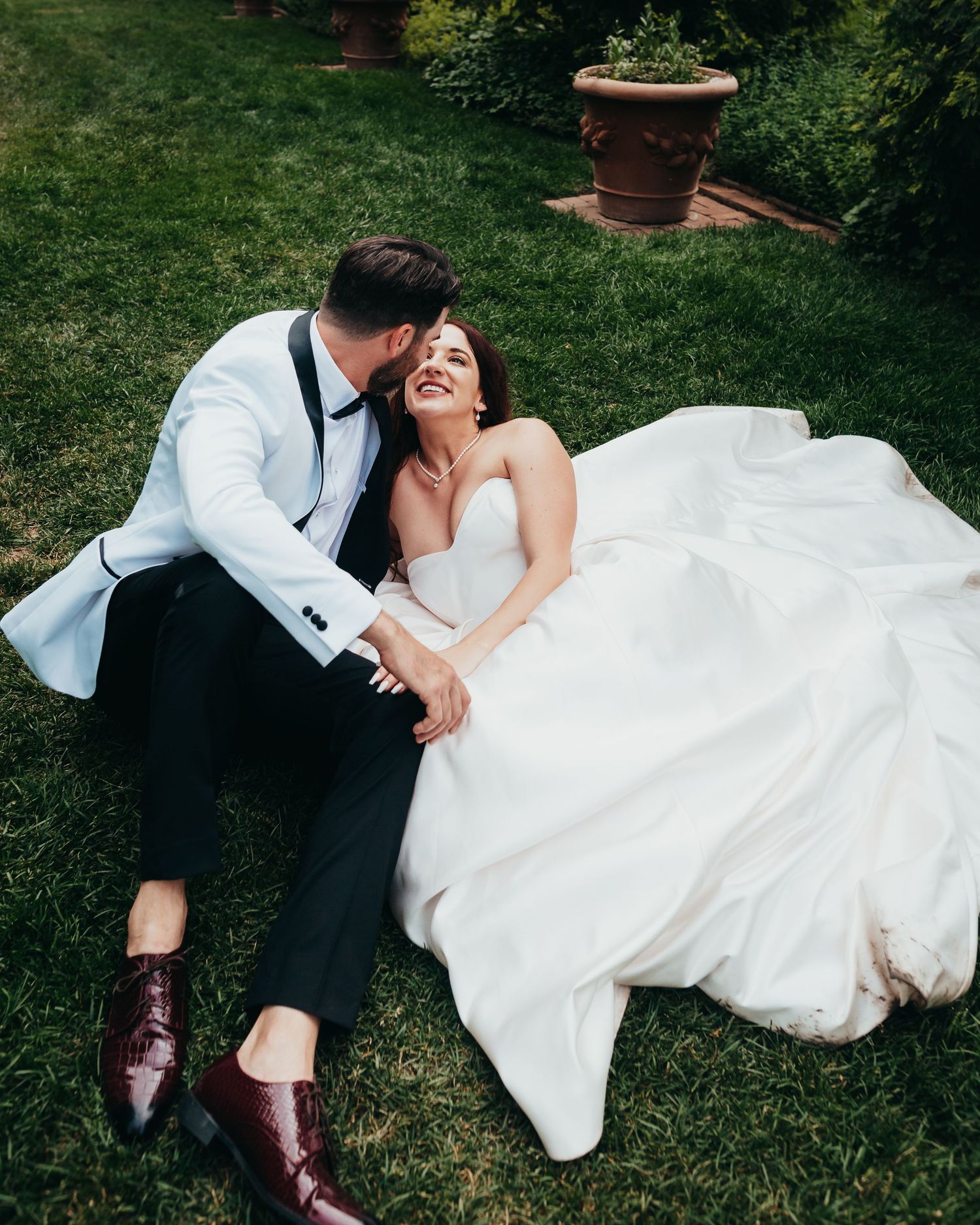 Newlyweds share intimate moment while laying on grass in garden setting.